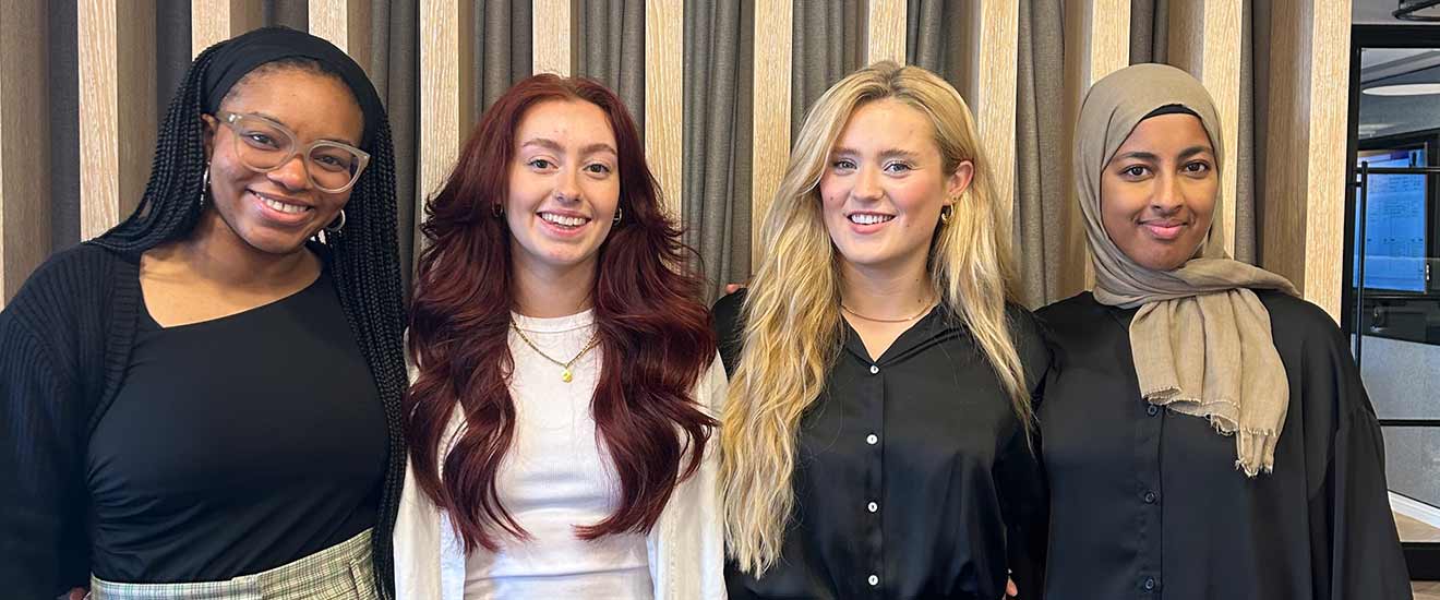 The image shows four interior design interns standing in front of a wooden joinery wall