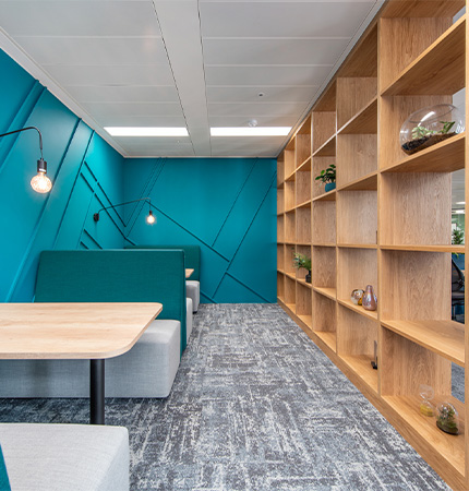 Modern office lounge with teal walls, wooden bookshelves, gray carpet, and comfortable seating under hanging lights.