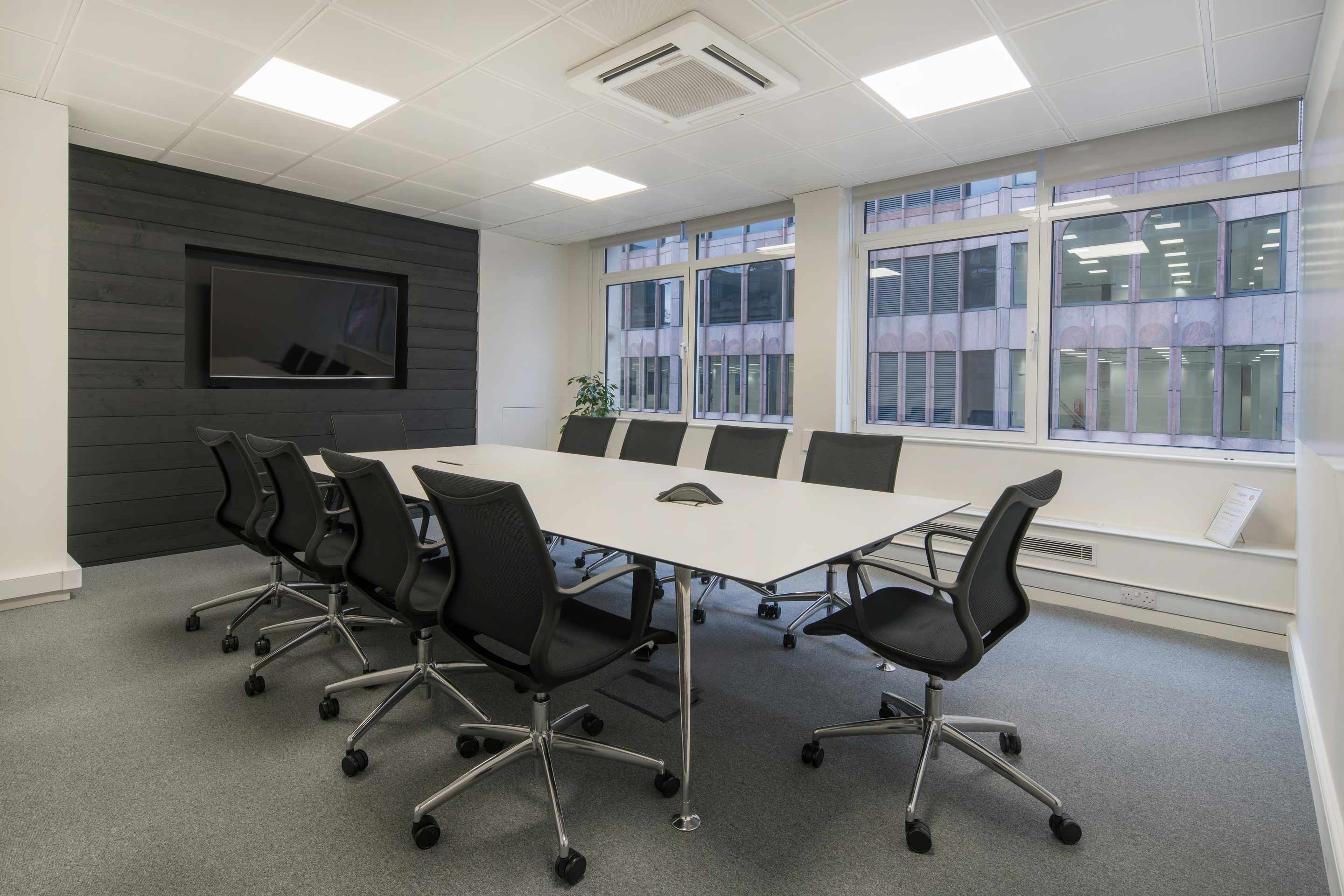 a large white meeting room with a grey carpet and a white rectangle table and a tv mounted on a black wall