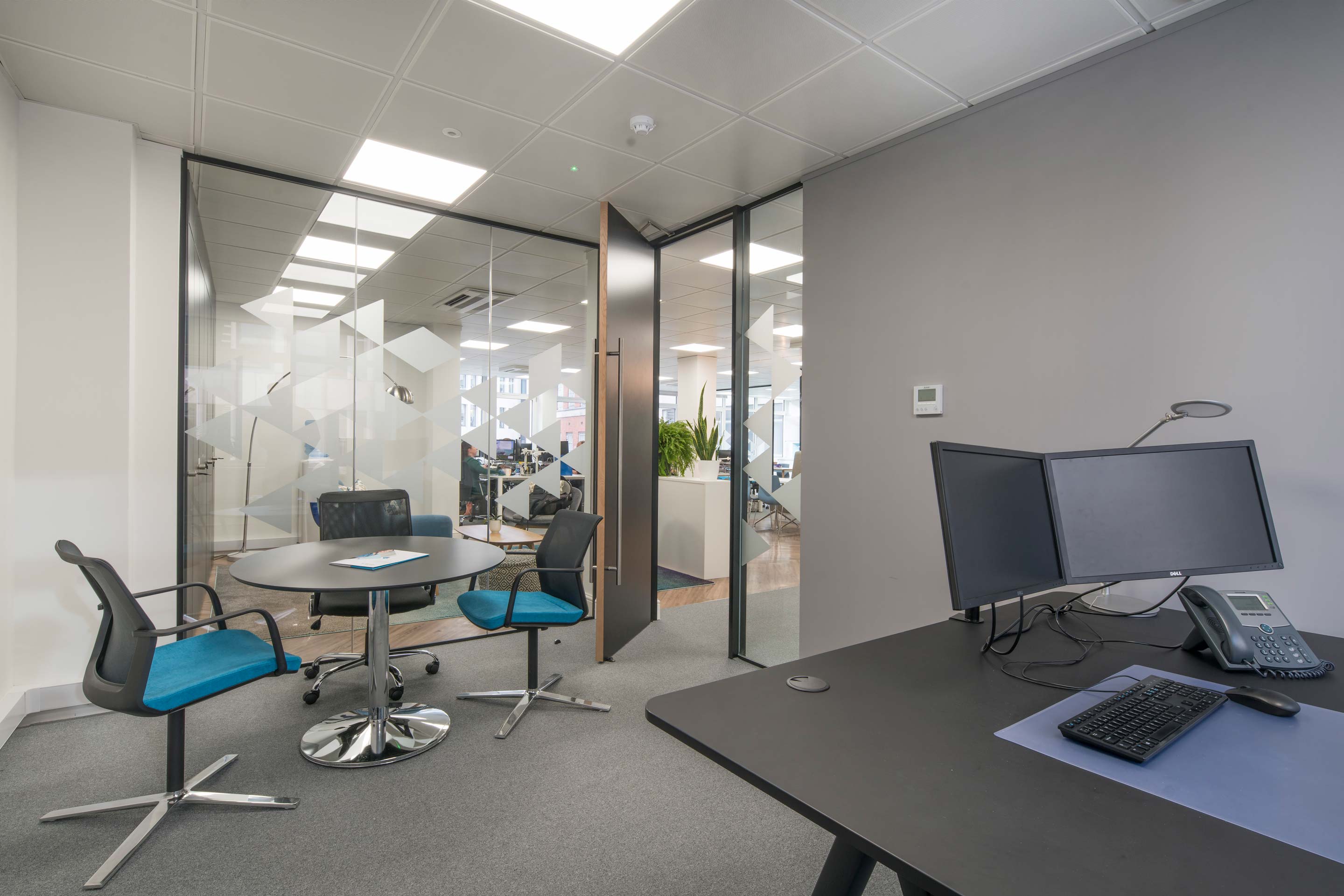 A grey and white office with a black desk on the right and a black circle table and seats on the left