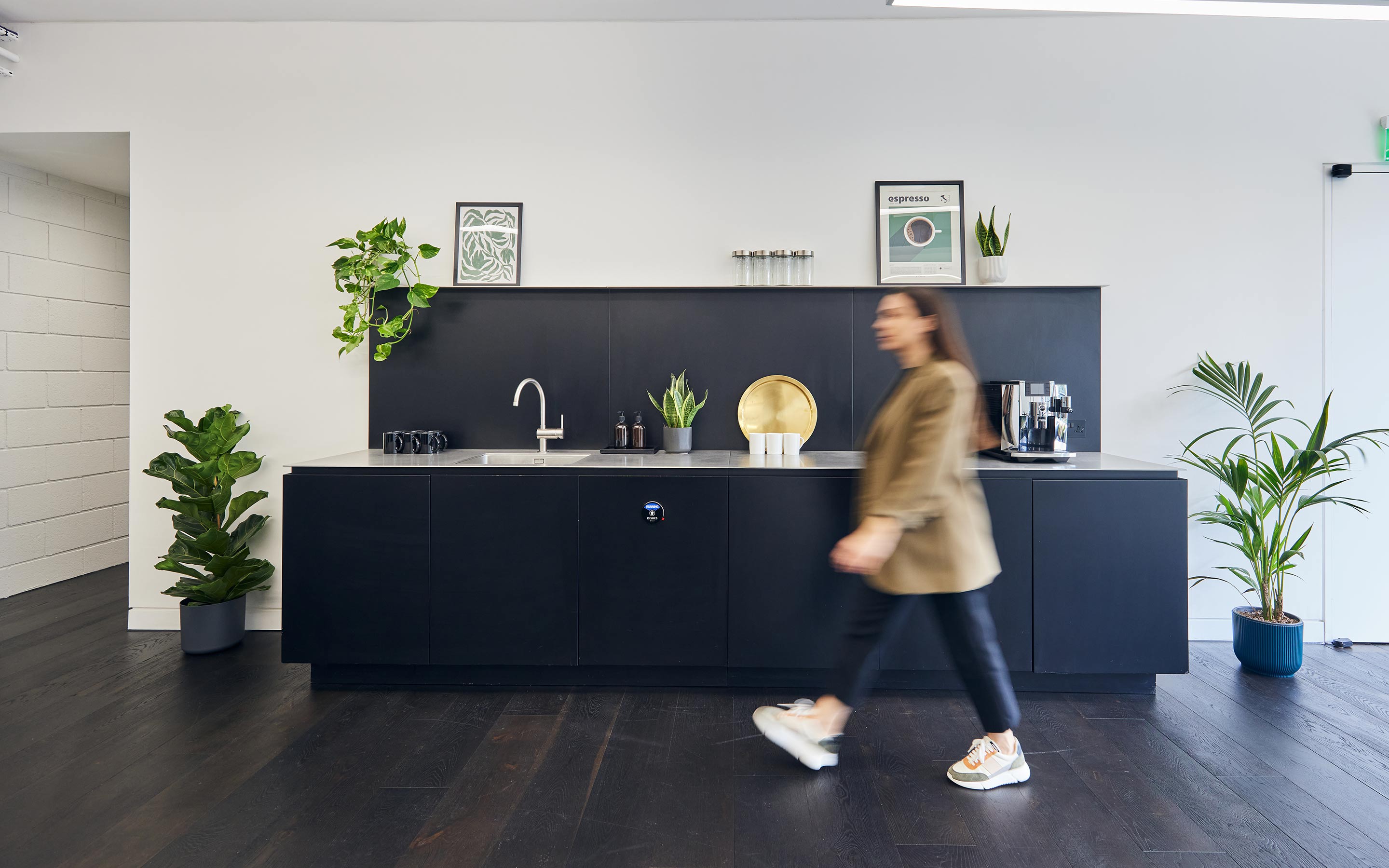 The image shows an employee walking through an office tea point. The space is complete with dark wooden flooring, black kitchen cabinets, and details such as plants and gold serving trays