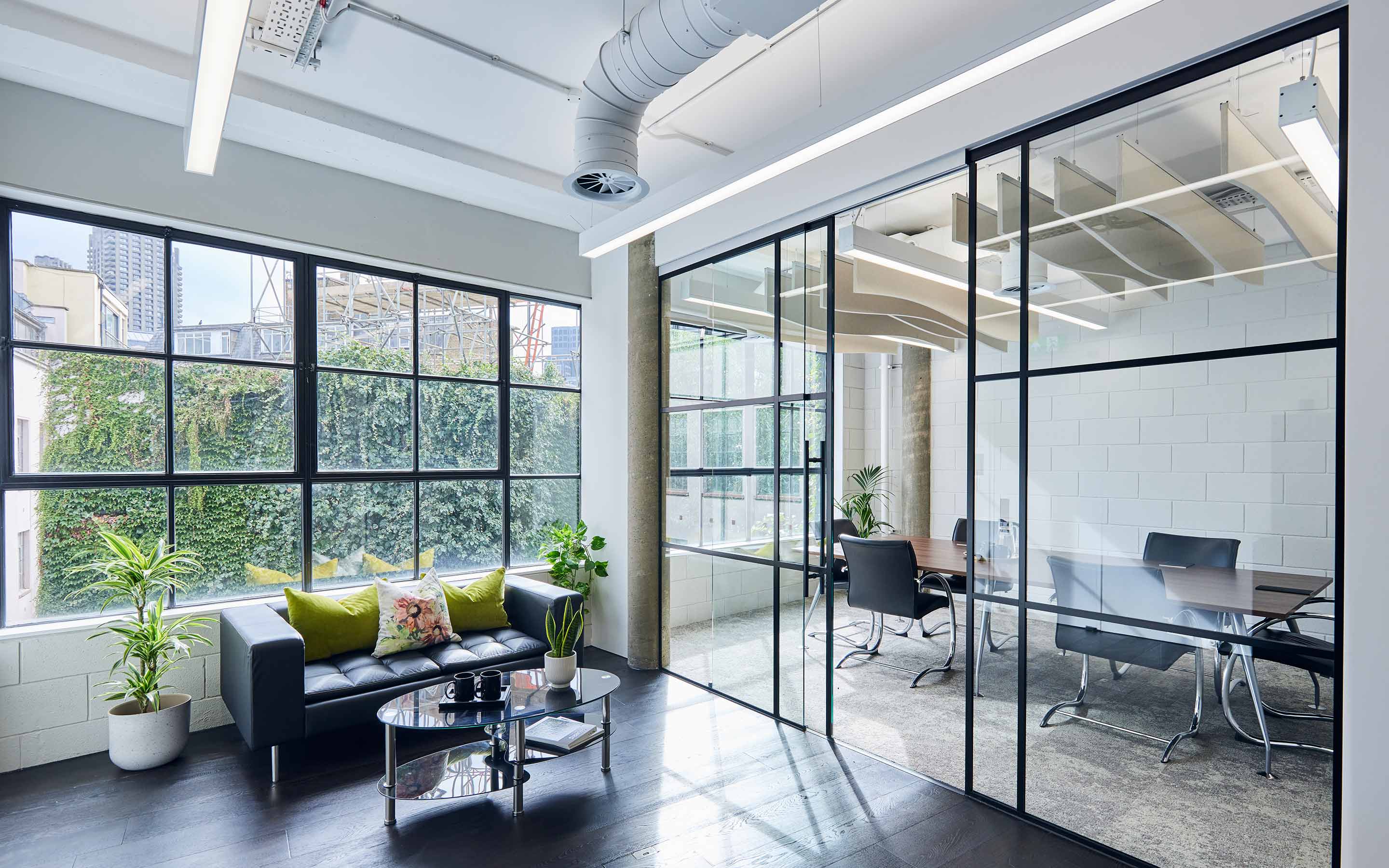 The image shows a glass-walled meeting room and a soft seating area in an office flooded with natural light