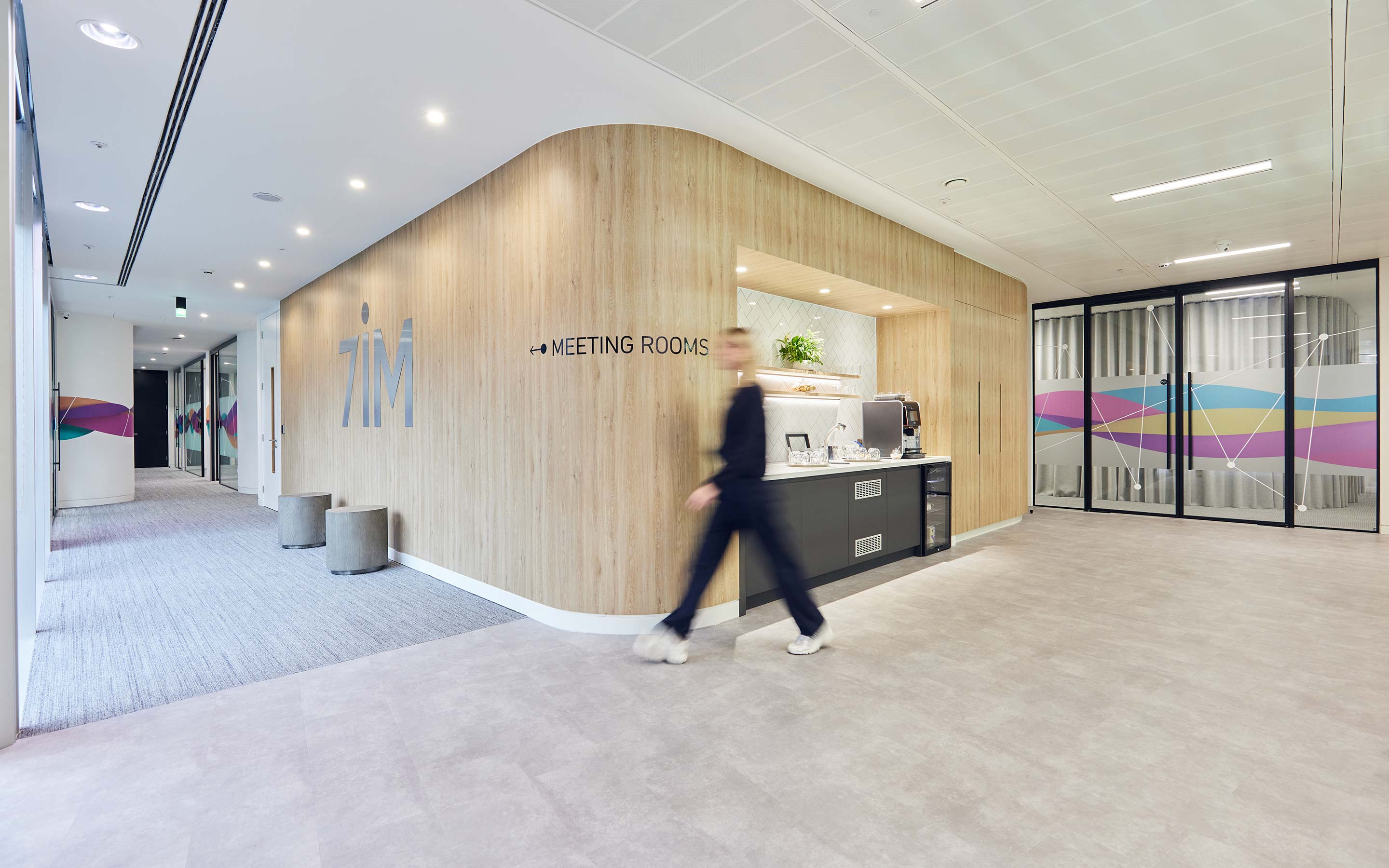 The image shows someone walking through a branded client reception area, complete with a teapoint, wooden wall panelling and soft seating