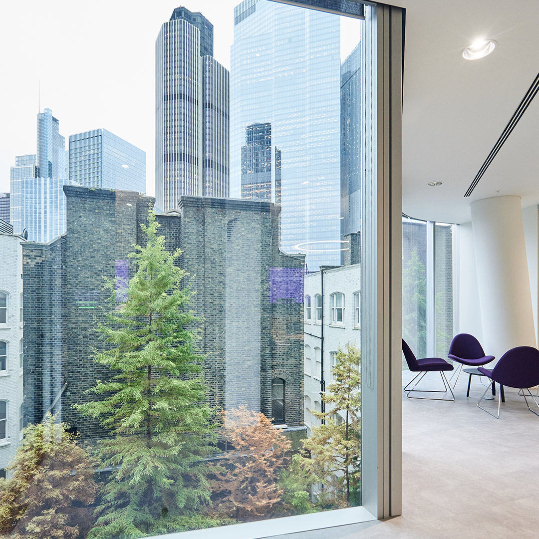 The image is of the city of London through a large office window. Soft seating in a purple colour is visible in the image's periphery.