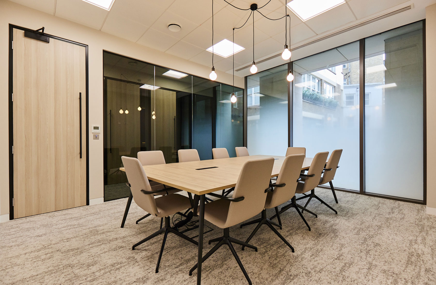 Feature lighting with exposed light bulbs hangs over a meeting room table with neutral coloured chairs and glass walls
