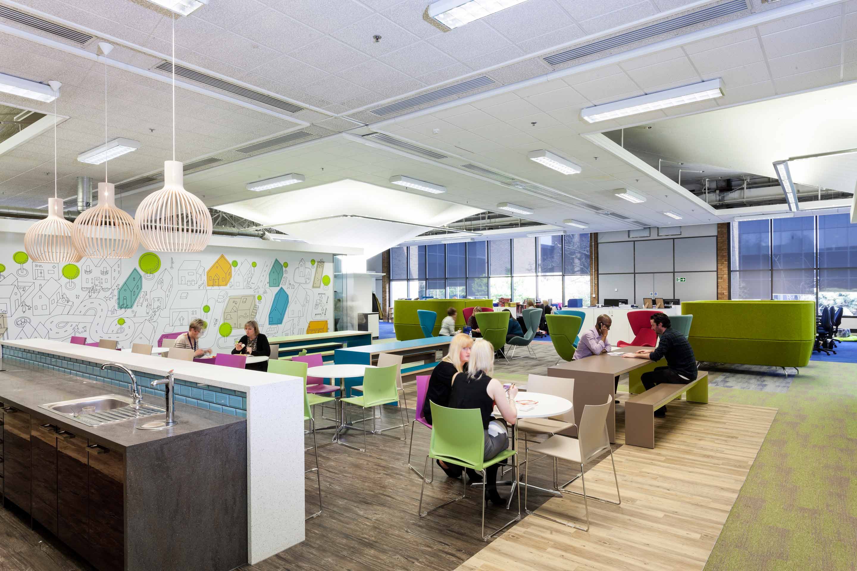 a dark stone effect kitchen space to the front left with a communal eating area with multicoloured chairs and benches with people eating lunch