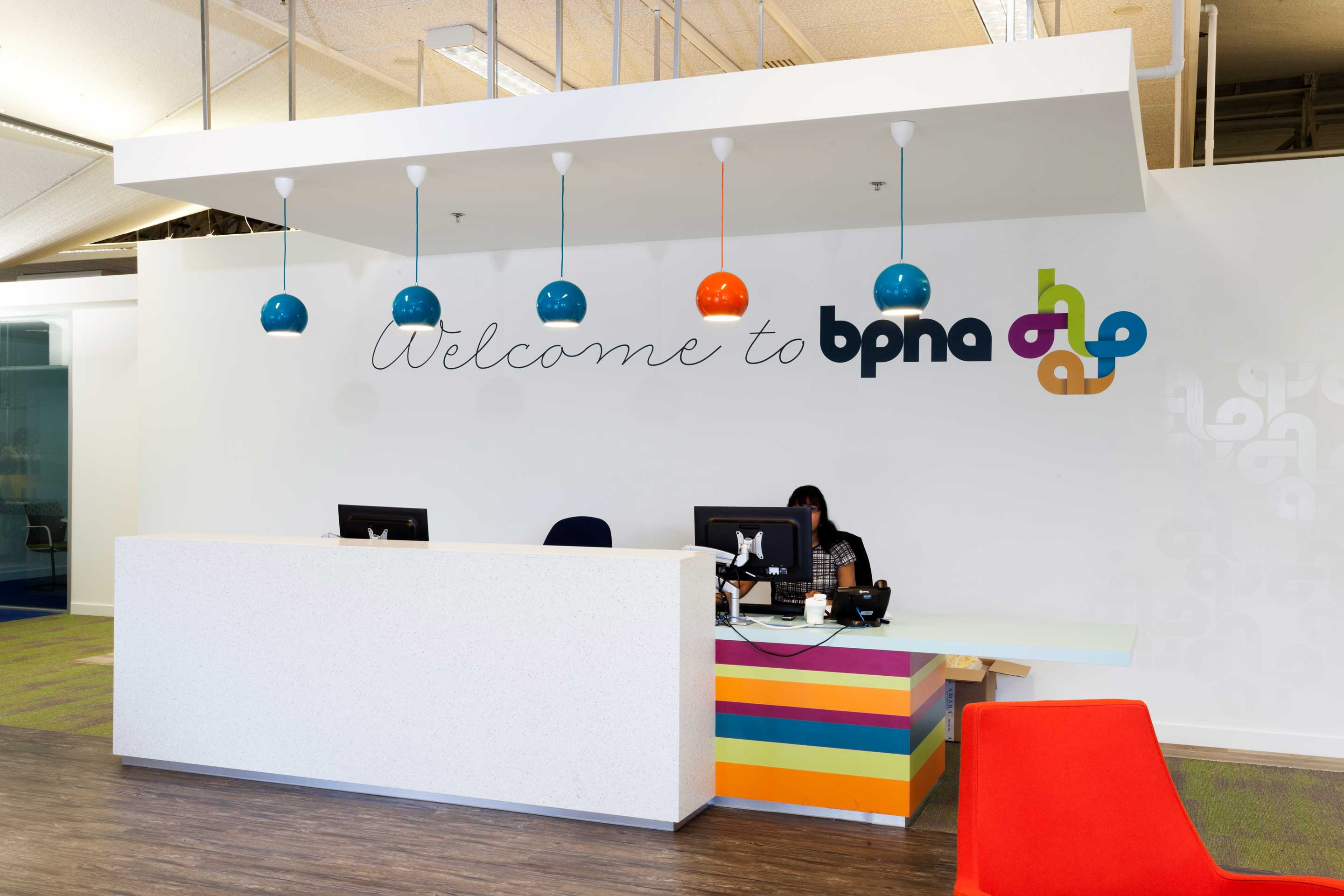 women sitting behind a white speckled front desk with low hanging blue and orange lighting on the right hand side of the front desk and a splash of multicoloured stripes