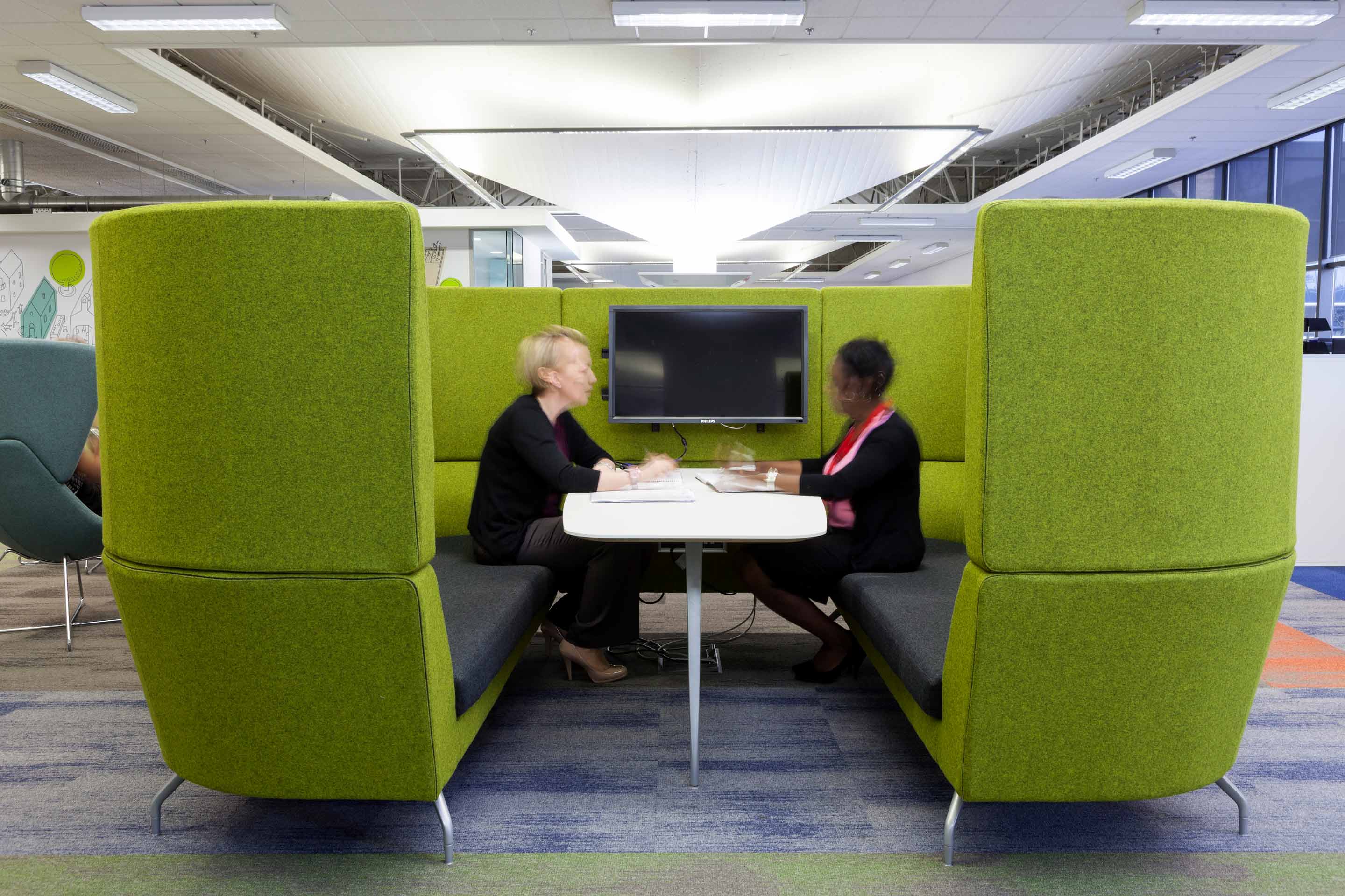 Two women sit across from one other in a green fabric high-backed sofa with a TV mounted in the centre.