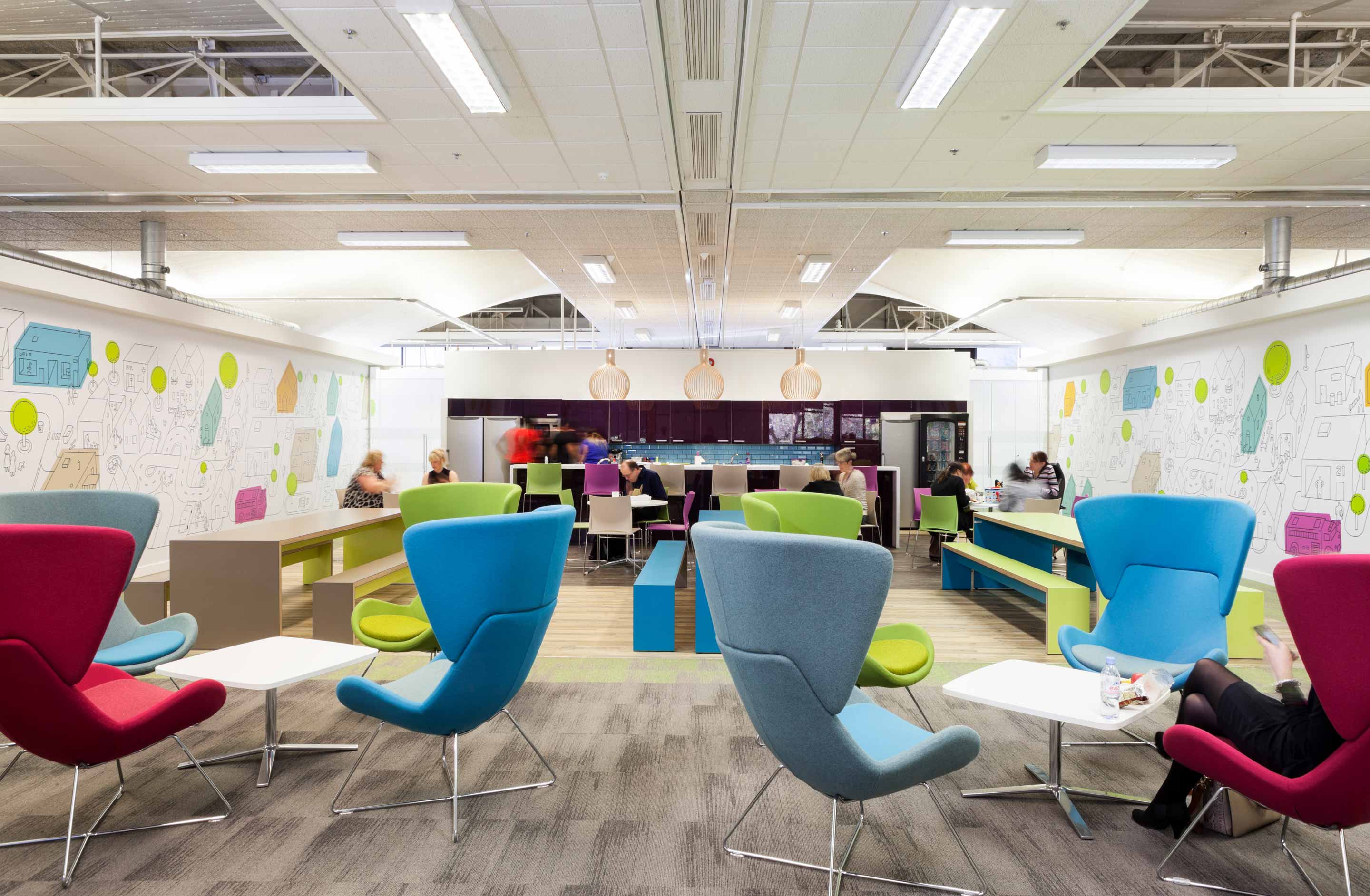 Purple kitchen area across the back with people eating food at tables and chairs in front with multi coloured high backed seats facing towards the kitchen area