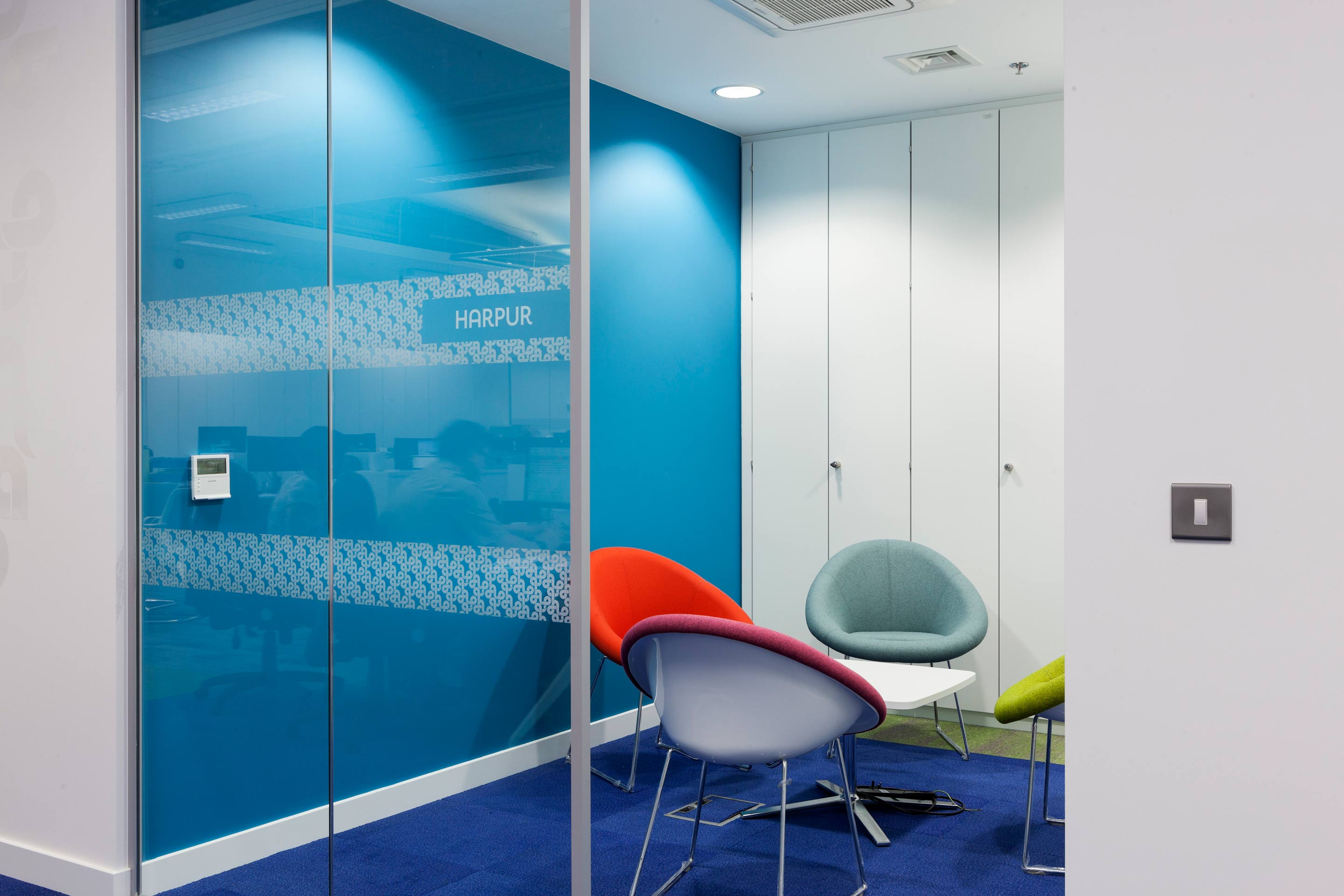 A blue meeting booth with multicoloured tub chairs facing inwards to a small white coffee table with a glass door.