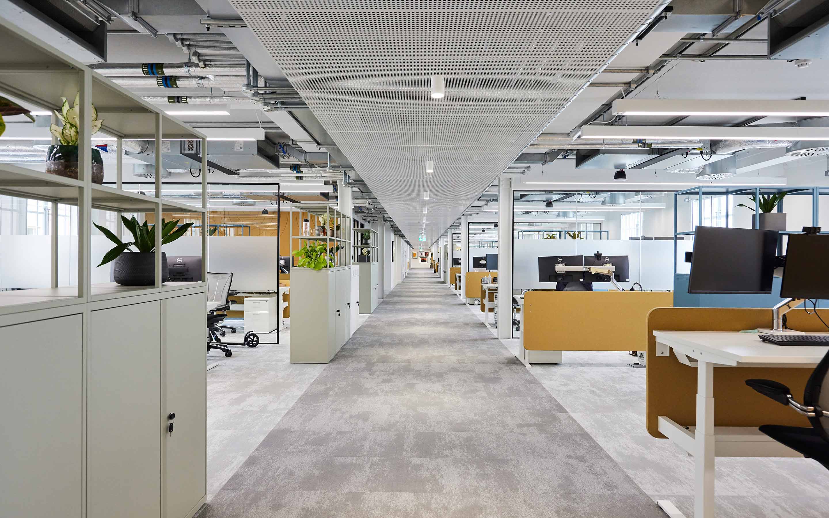The corridor through an open plan office, with desks, acoustic panels, exposed mechanical and engineering services, and a carpeted floor