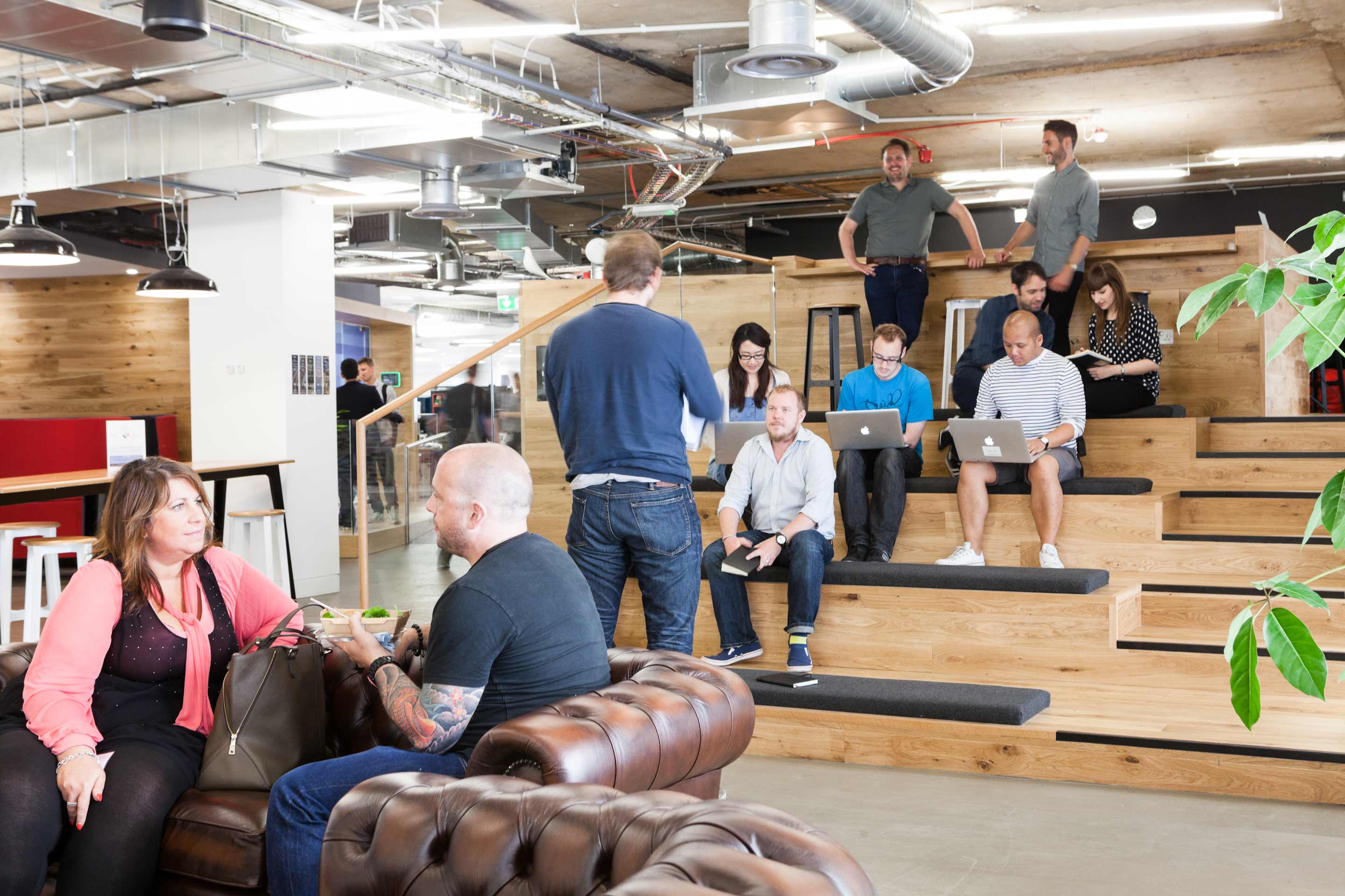 In the centre of the photograph a group of individuals are seated on a wooden staircase seating area with two people in front seated on brown leather sofas