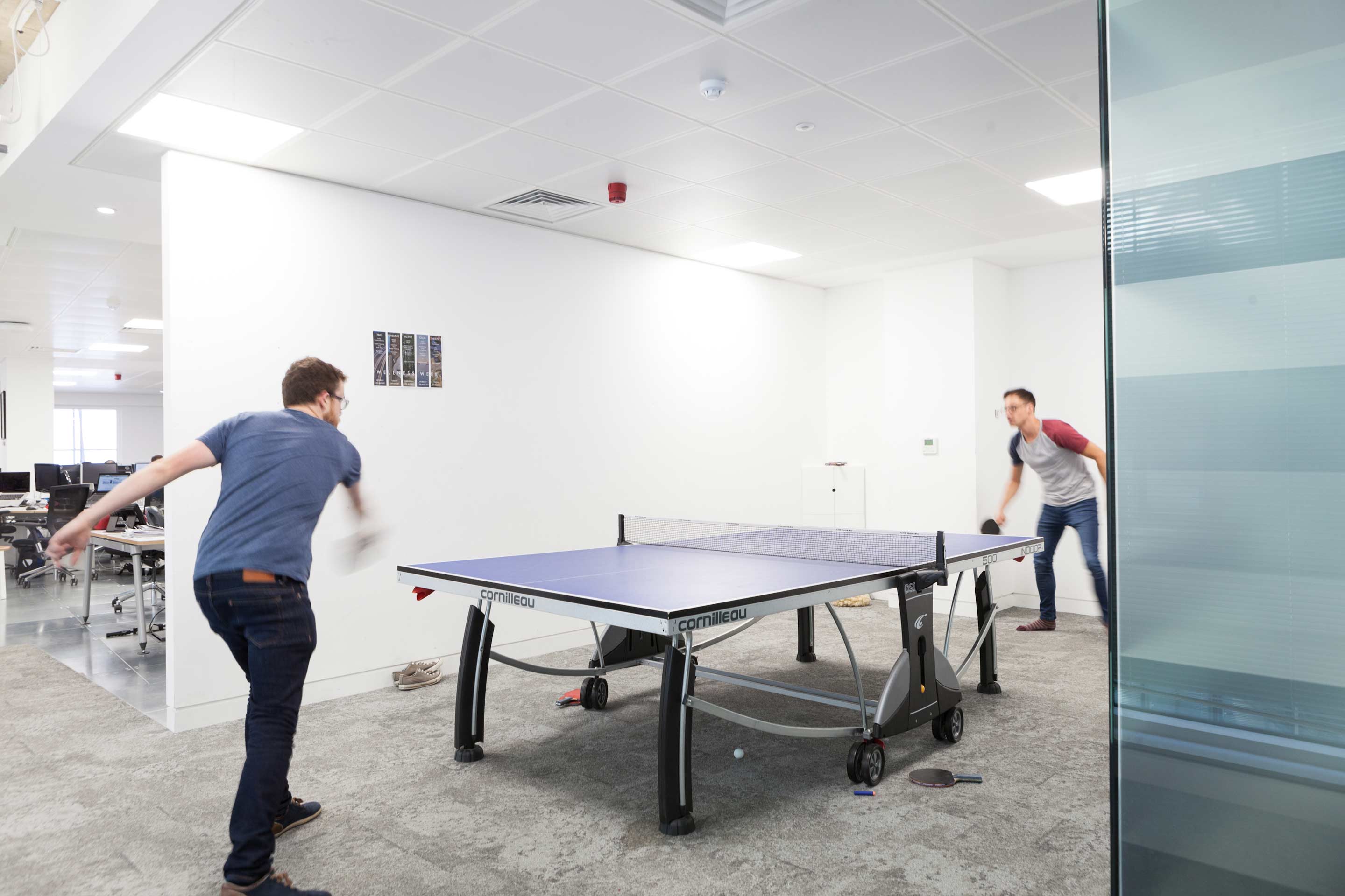 A blue table tennis table in the centre  with two people holding bats at either end surrounded by white and glass walls