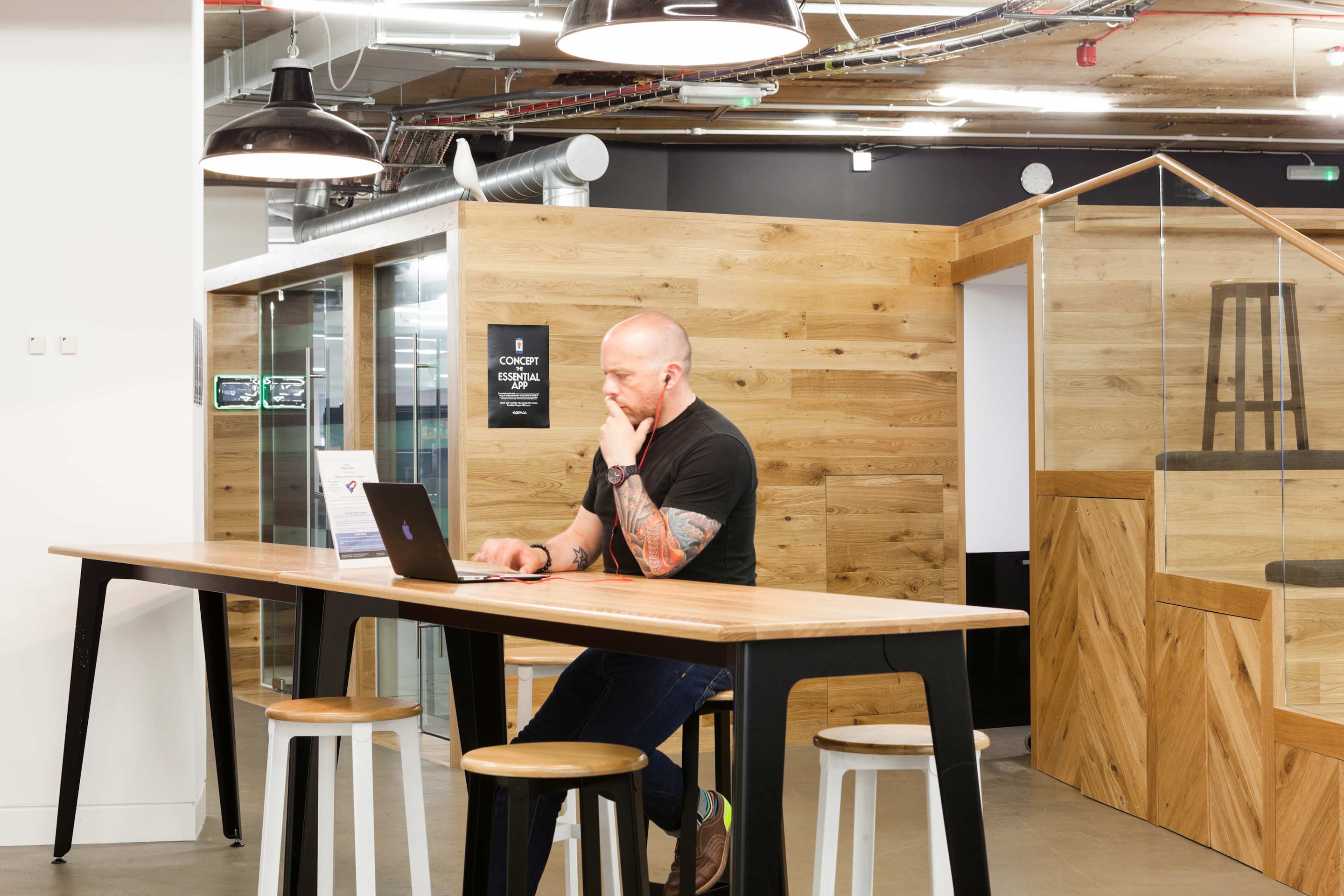 A man sat at a breakfast bar-style wooden table with a black laptop surrounded by wooden pods and a seating staircase space on the right