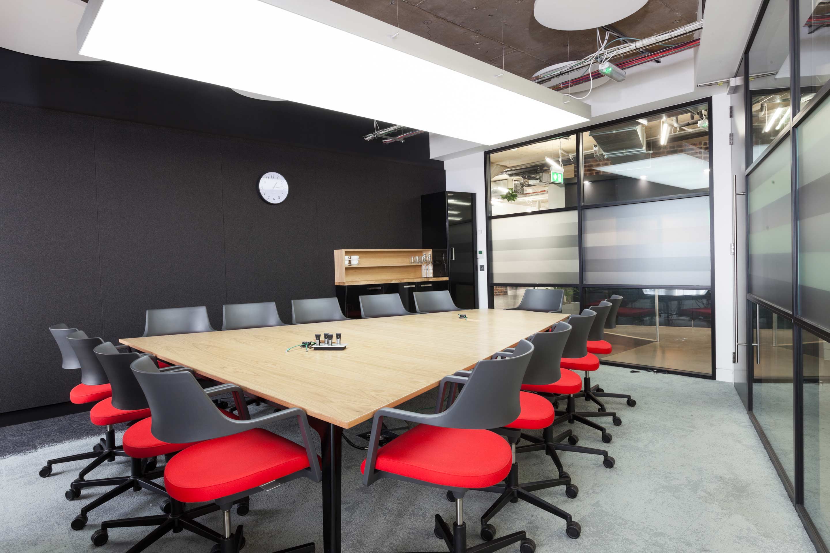 A light hardwood Isoosceles trapezoid boardroom table with black and red desk chairs in the centre of a black and glass walled meeting room with a small wooden kitchen area in the farback wall