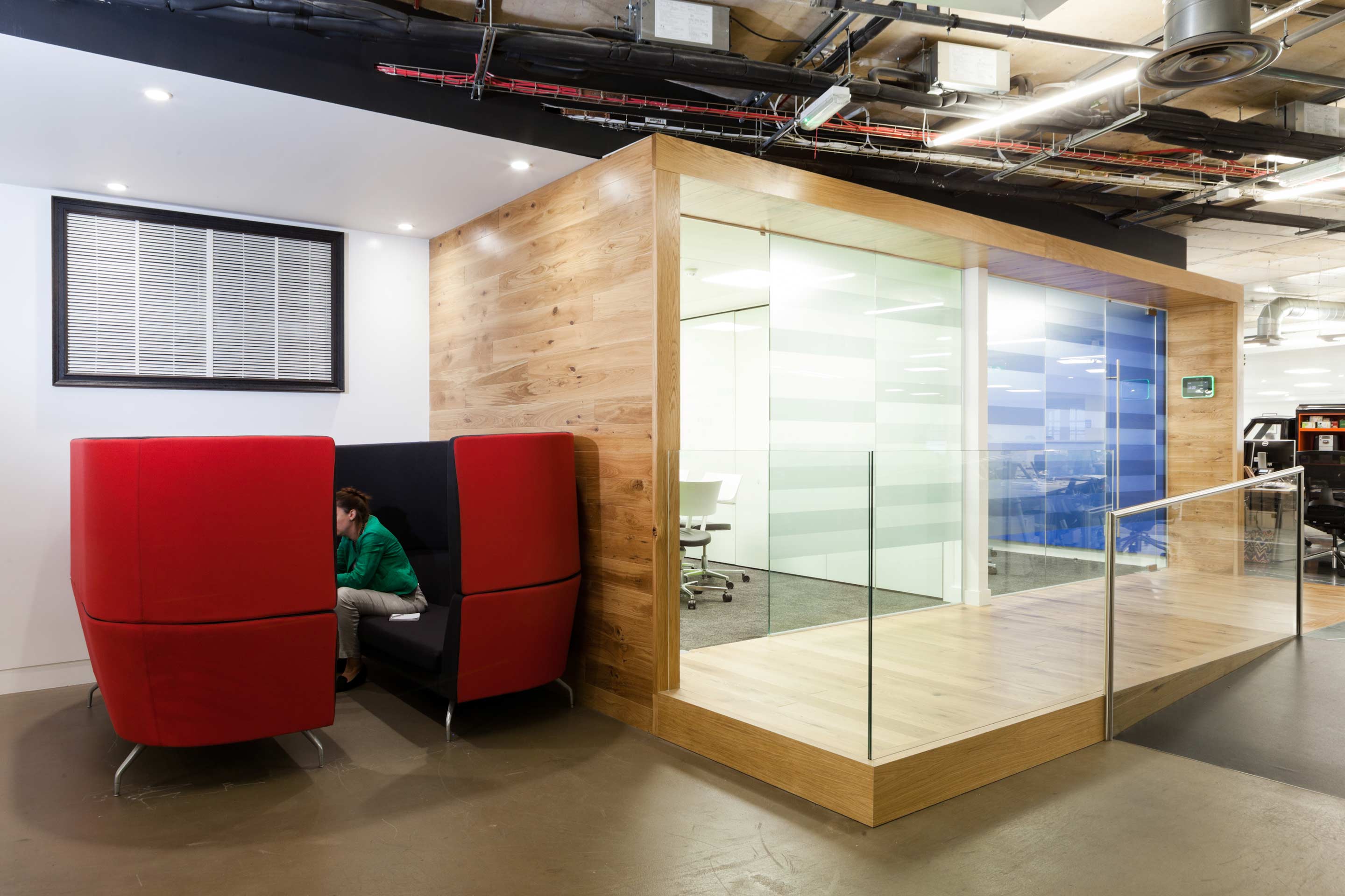 two wooden cube-shaped meeting booths with glass fronts positioned on a wooden ramp to the right, and two red high-backed sofas