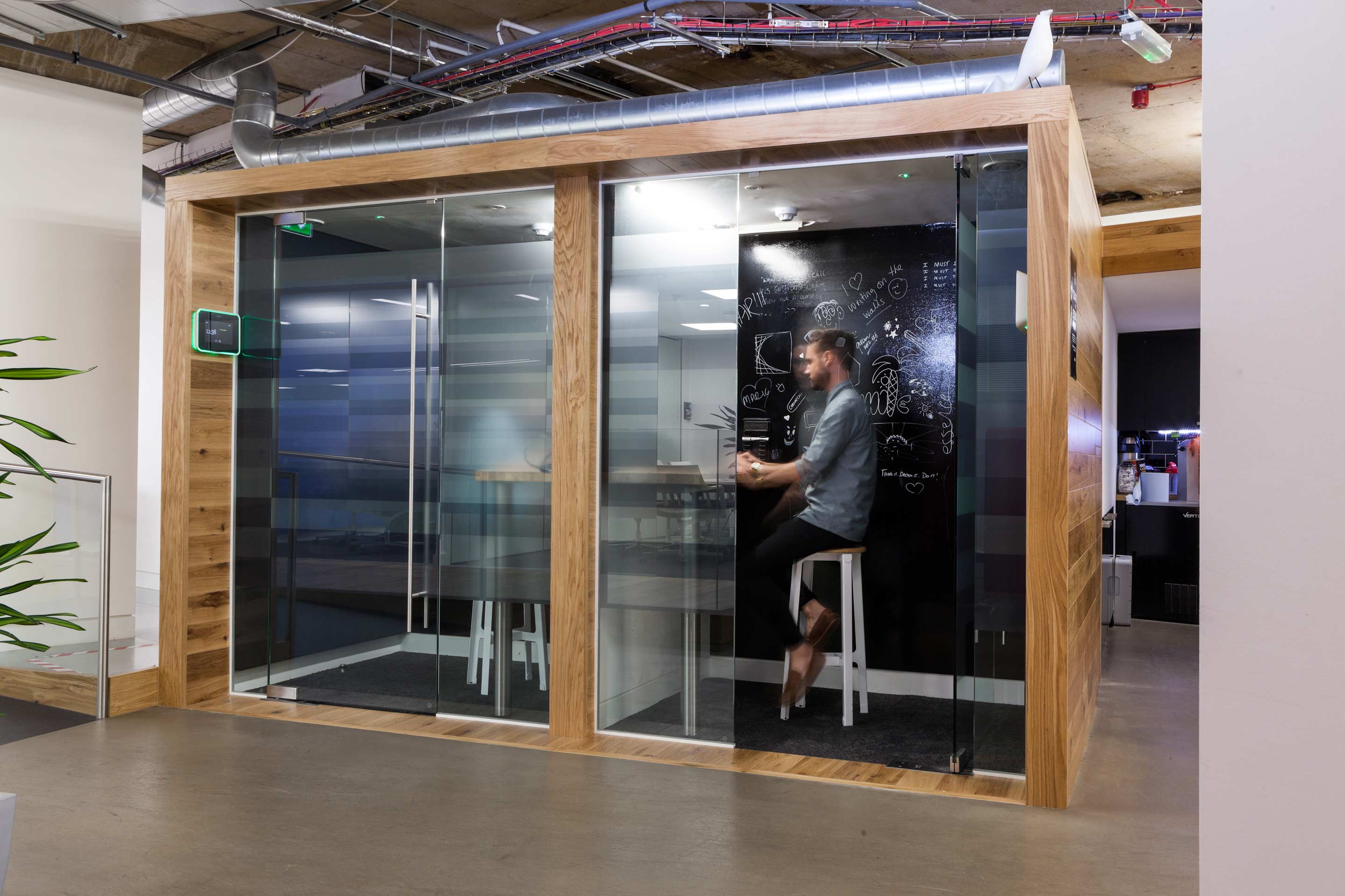 two wooden pods with glass doors and white chairs and a man seated in the right hand pod with a black white board with white lettering