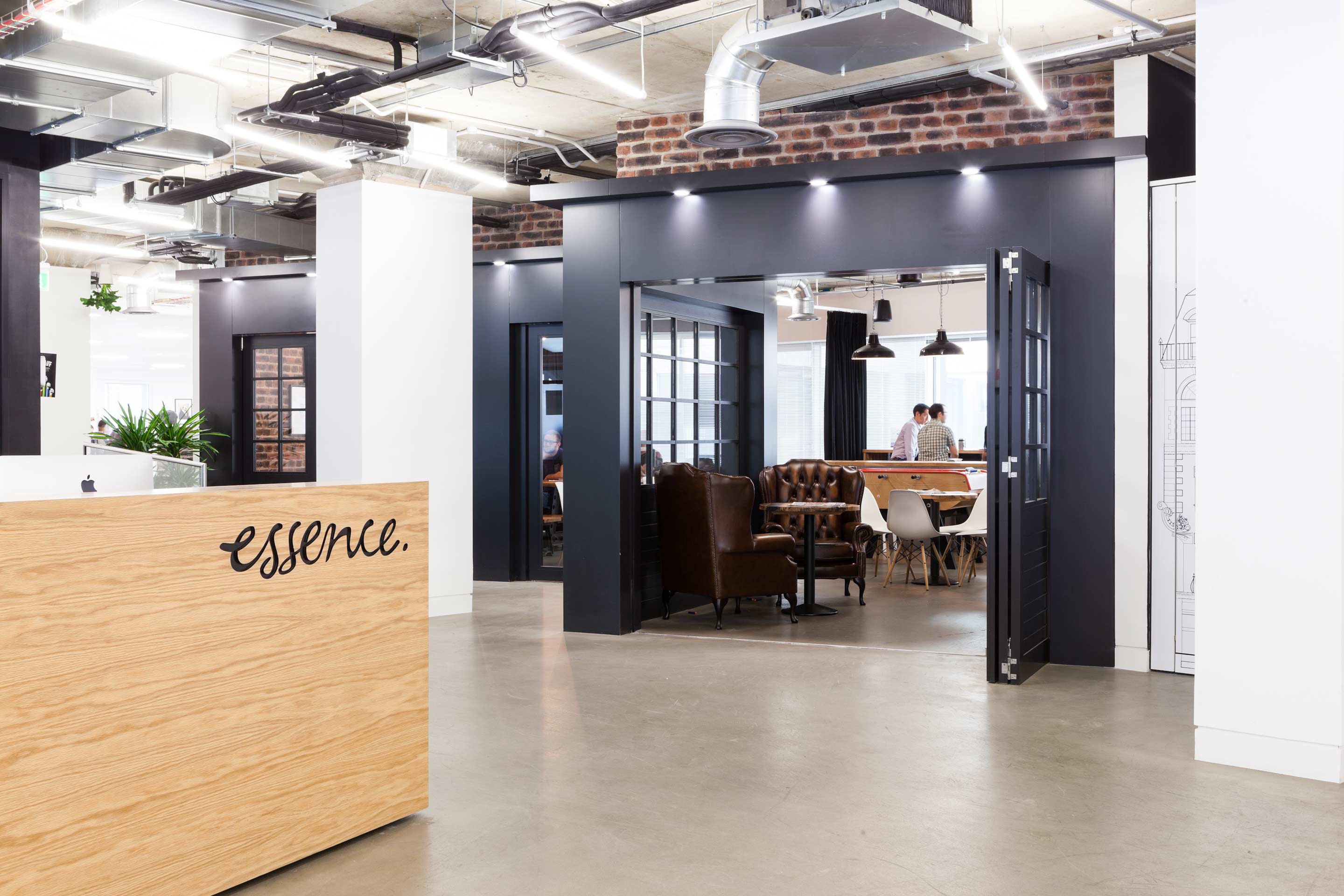 On the left is a light wooden effect front desk with a black cube shaped communal room with brown leather chairs and two persons enjoying coffee