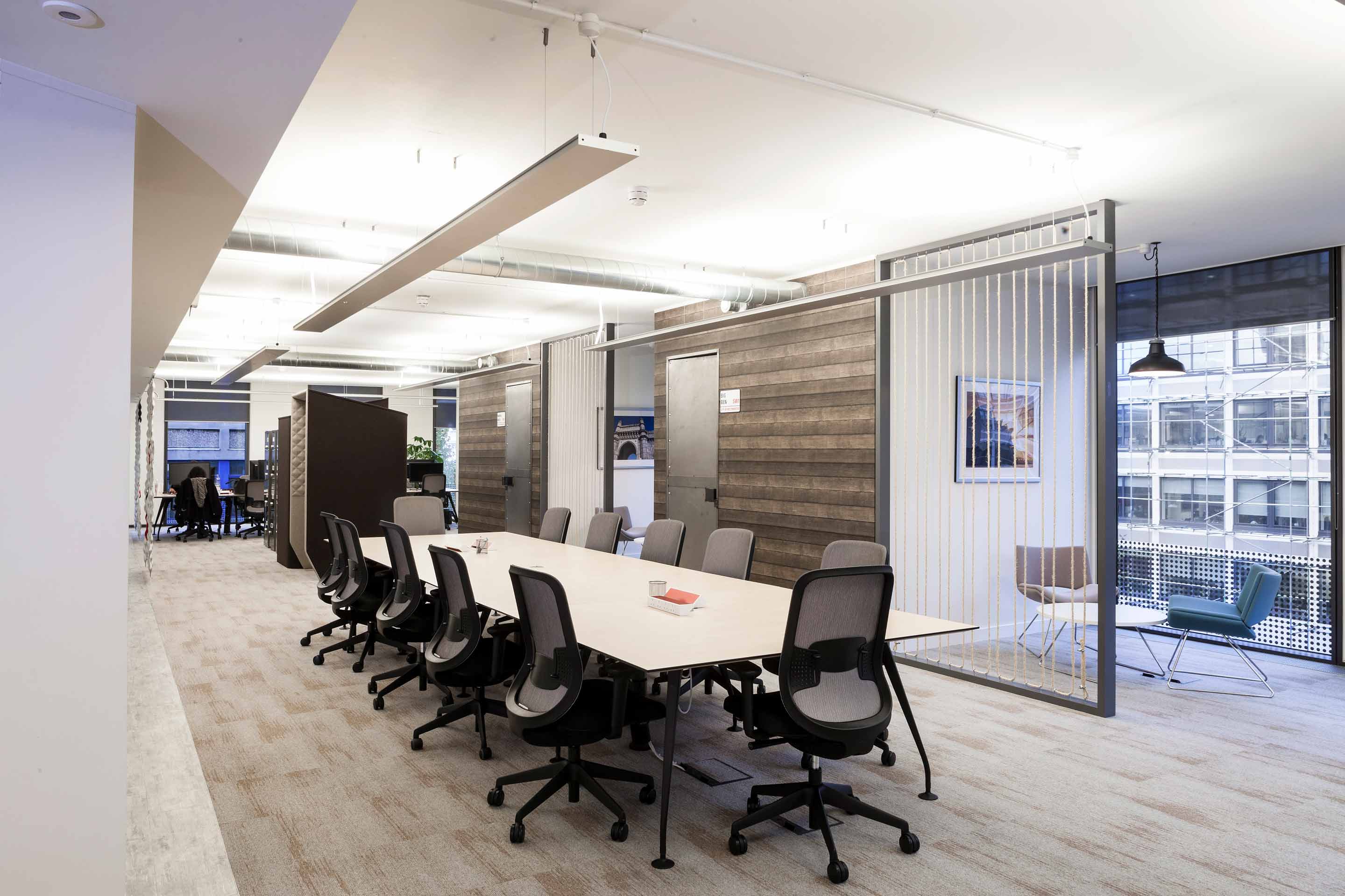 A white boardroom table with black seats in the centre of an open plan space with meeting rooms on the right.