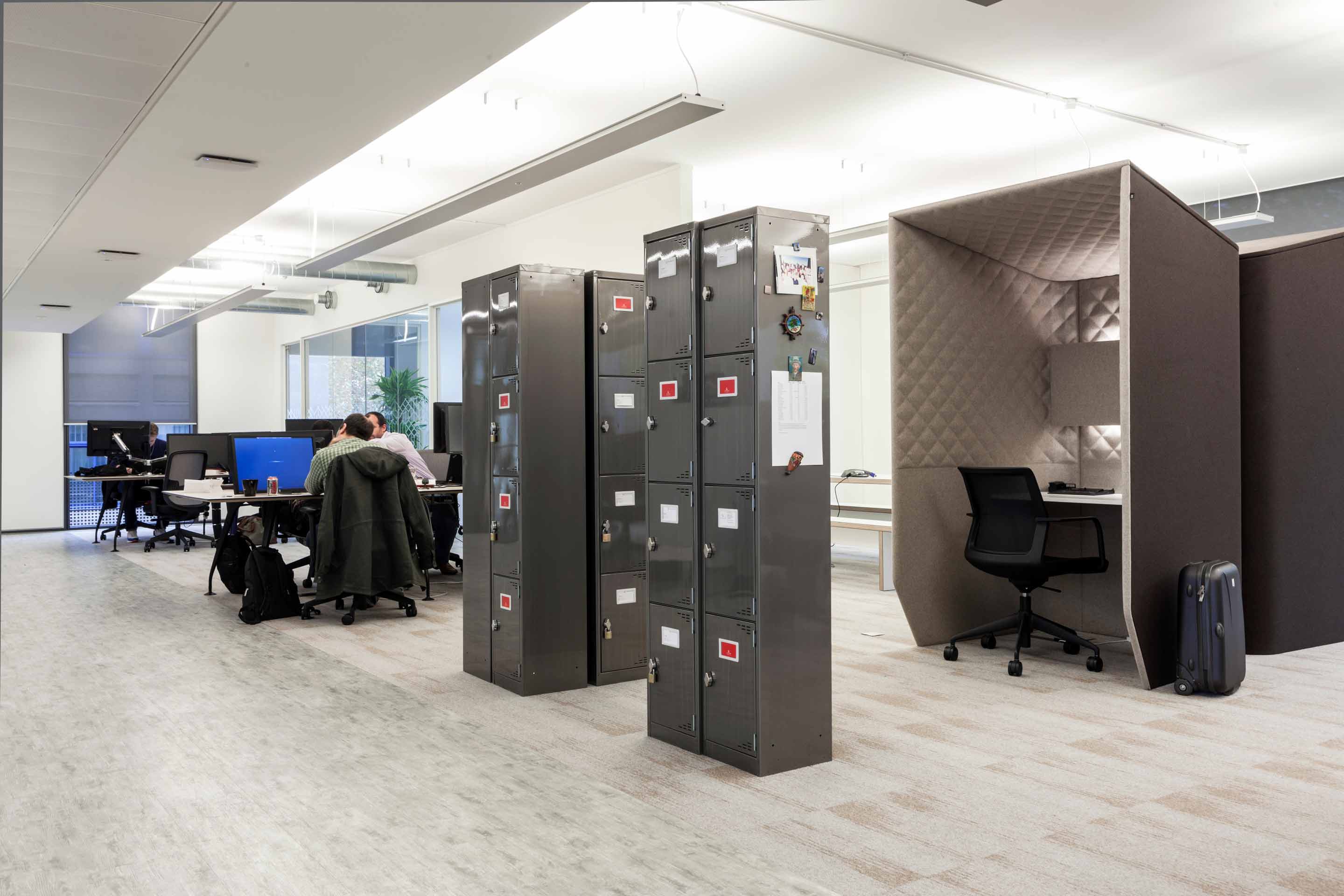 Black metal lockers located in the centre of an open office area with a fabric pod on the right and white desk spaces on the opposite side of the room