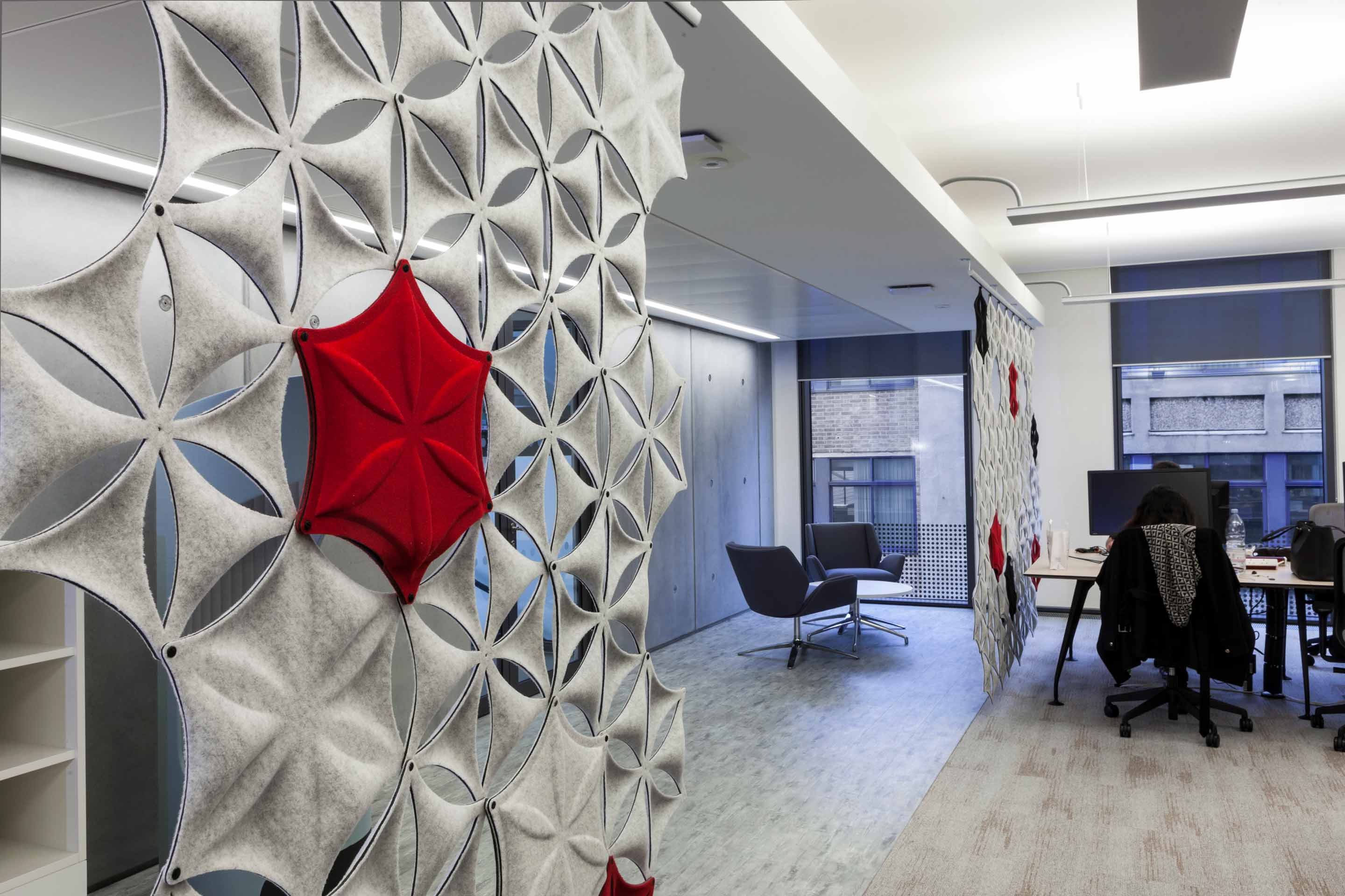 a light grey fabric wall panel with red accents separating a two-person meeting room and white desks with black chairs and monitors