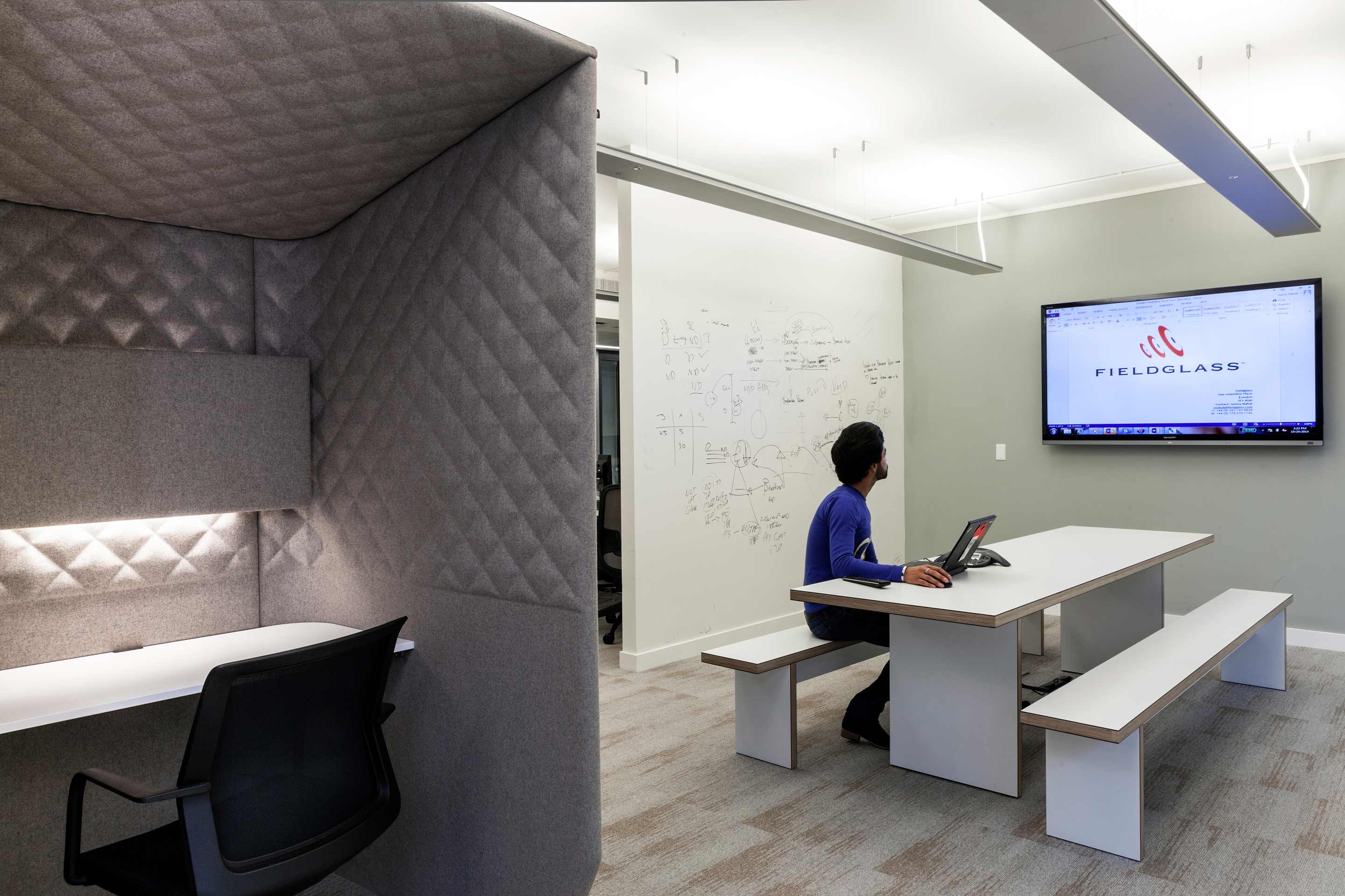 A white table and benches in front of a mounted TV screen with a white board wall and a grey fabric pod to the left