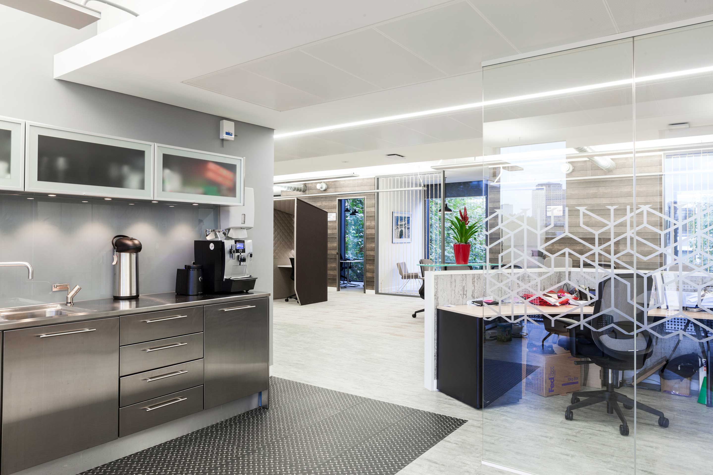 Metal kitchen on the left with a glass partition on the right to an open plan walk way and desk space