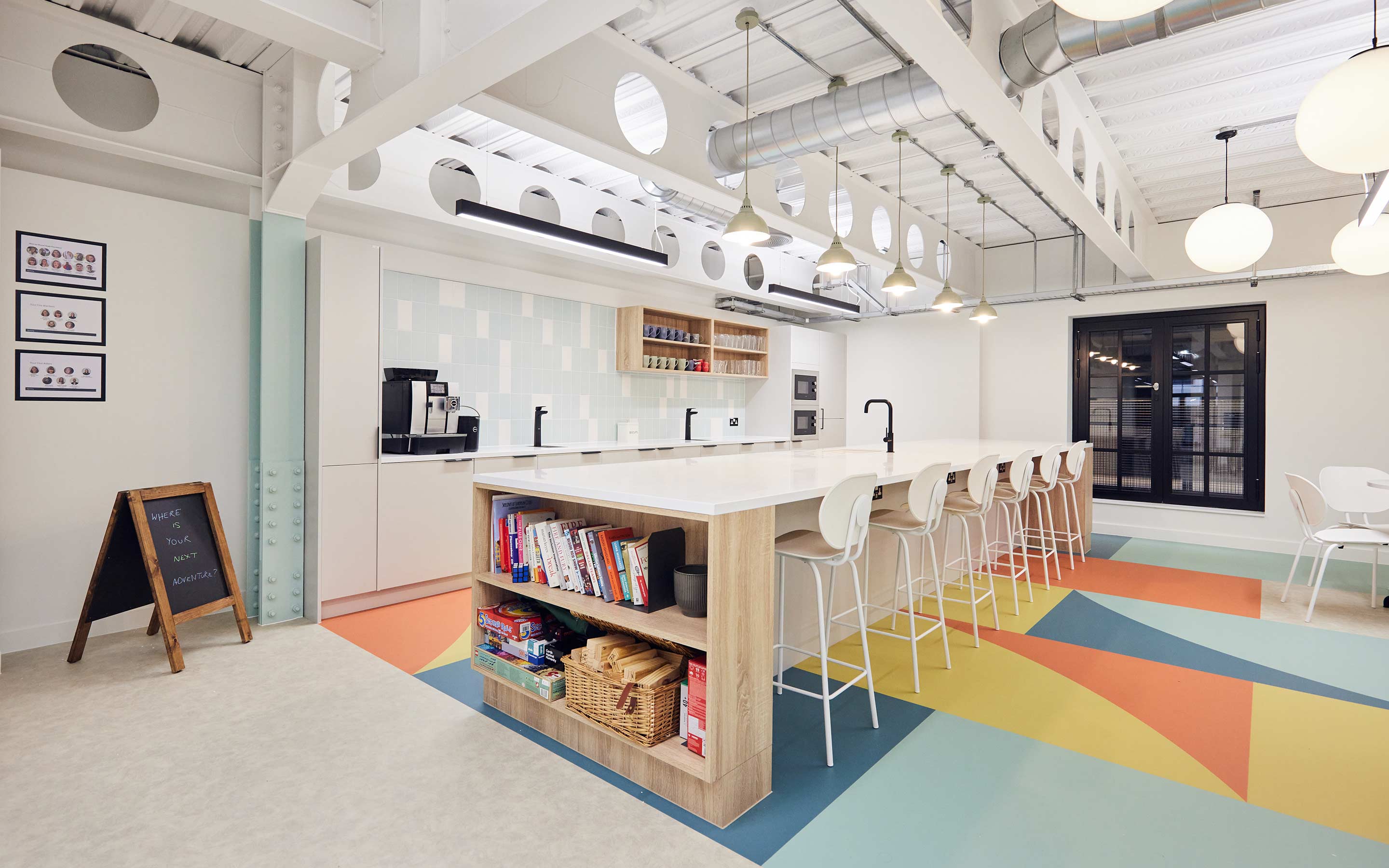 The image shows an office tea point area with colourful flooring, a breakfast bar with stools and sinks, and pendant lighting