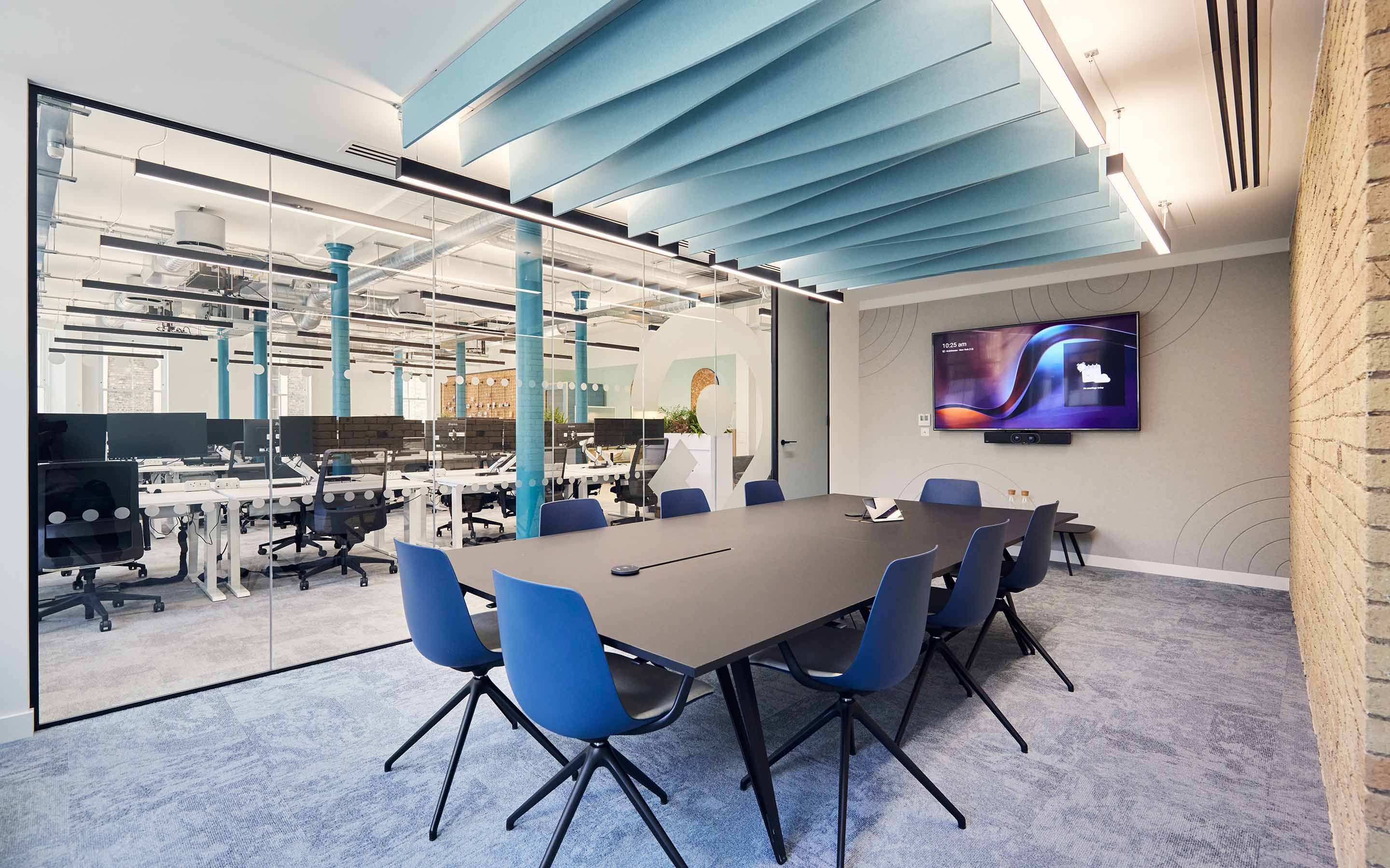 The image shows a boardroom with acoustic ceiling panels, an exposed brick wall, and views of the open-plan office beyond