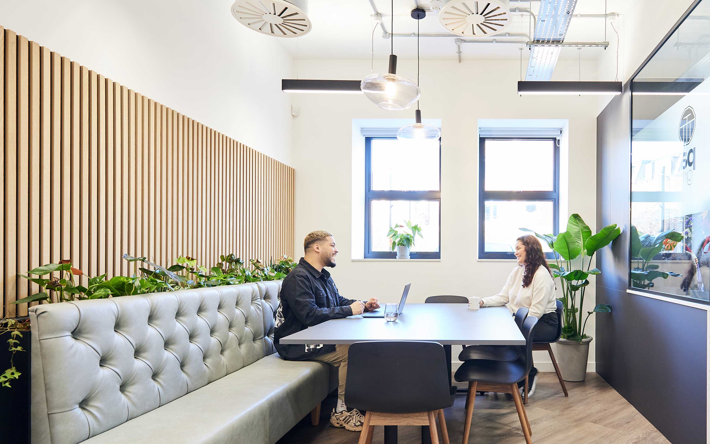 Office space with employees laughing at a stylish table and chairs, with lots of interior plants and exposed mechanical and electrical services