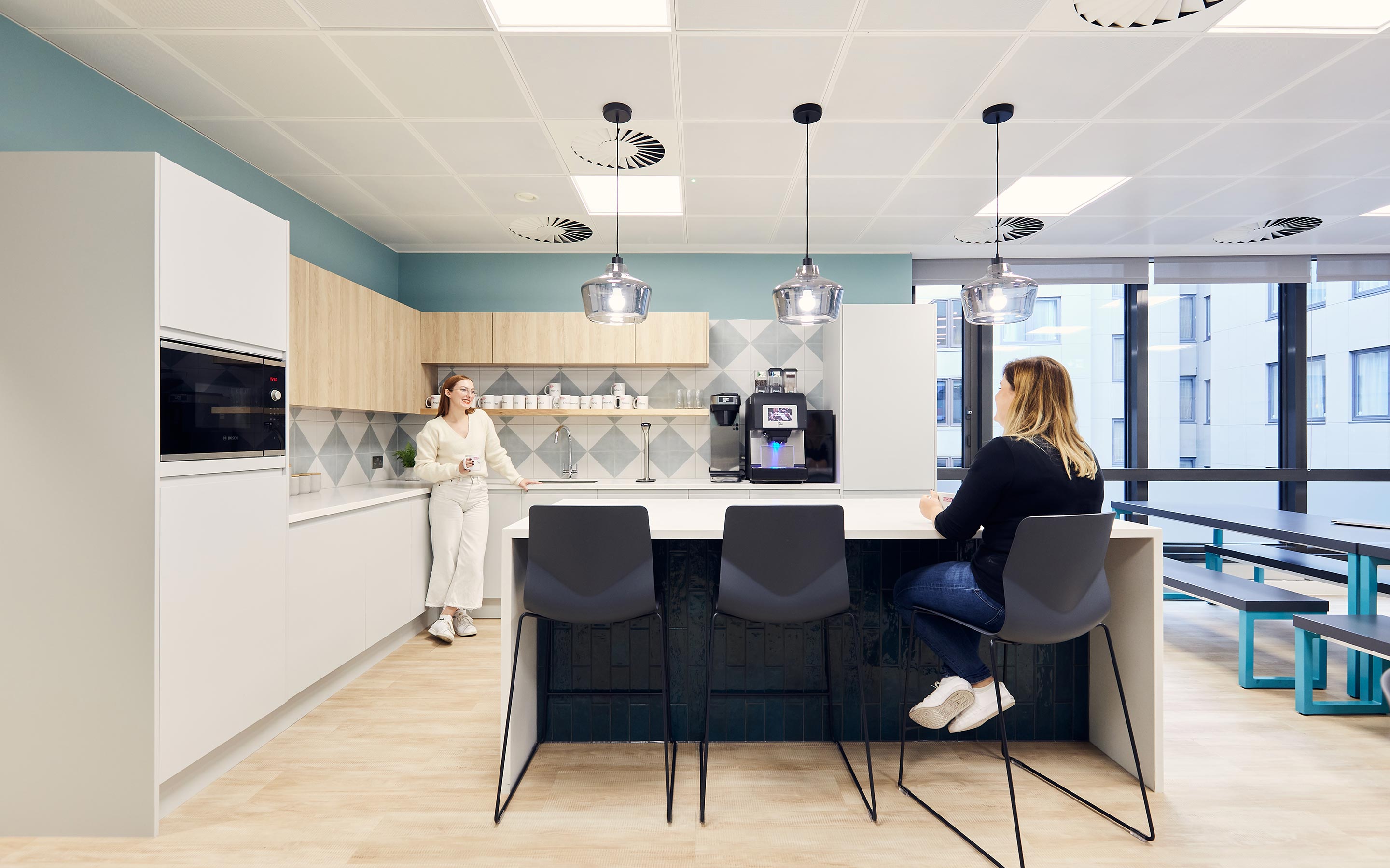 The images shows employees talking at an office kitchen counter. The aesthetic is bright, and welcoming