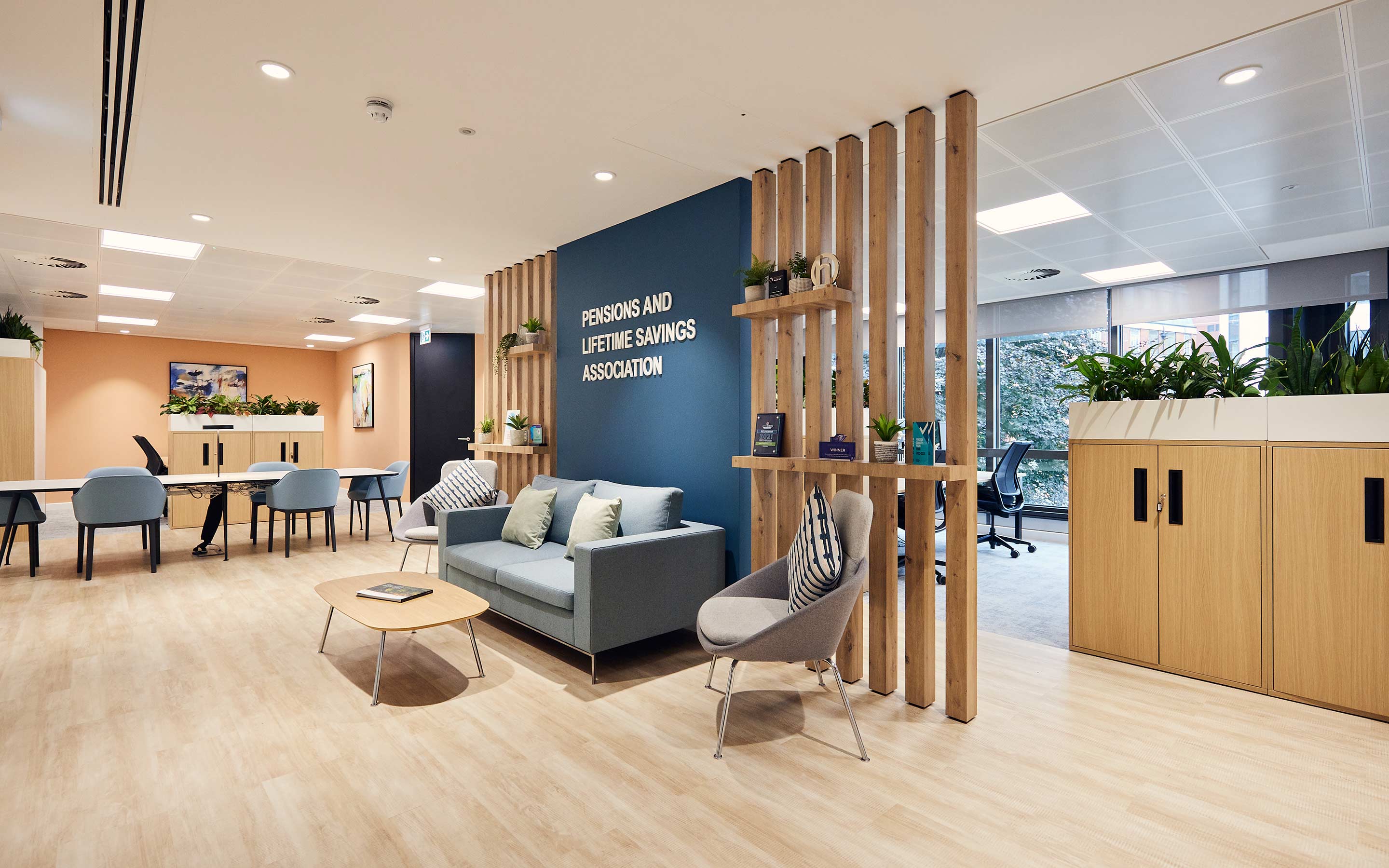 The images shows a client arrival area in an office, with soft seating, bespoke joinery, and elements of desking in the wider office