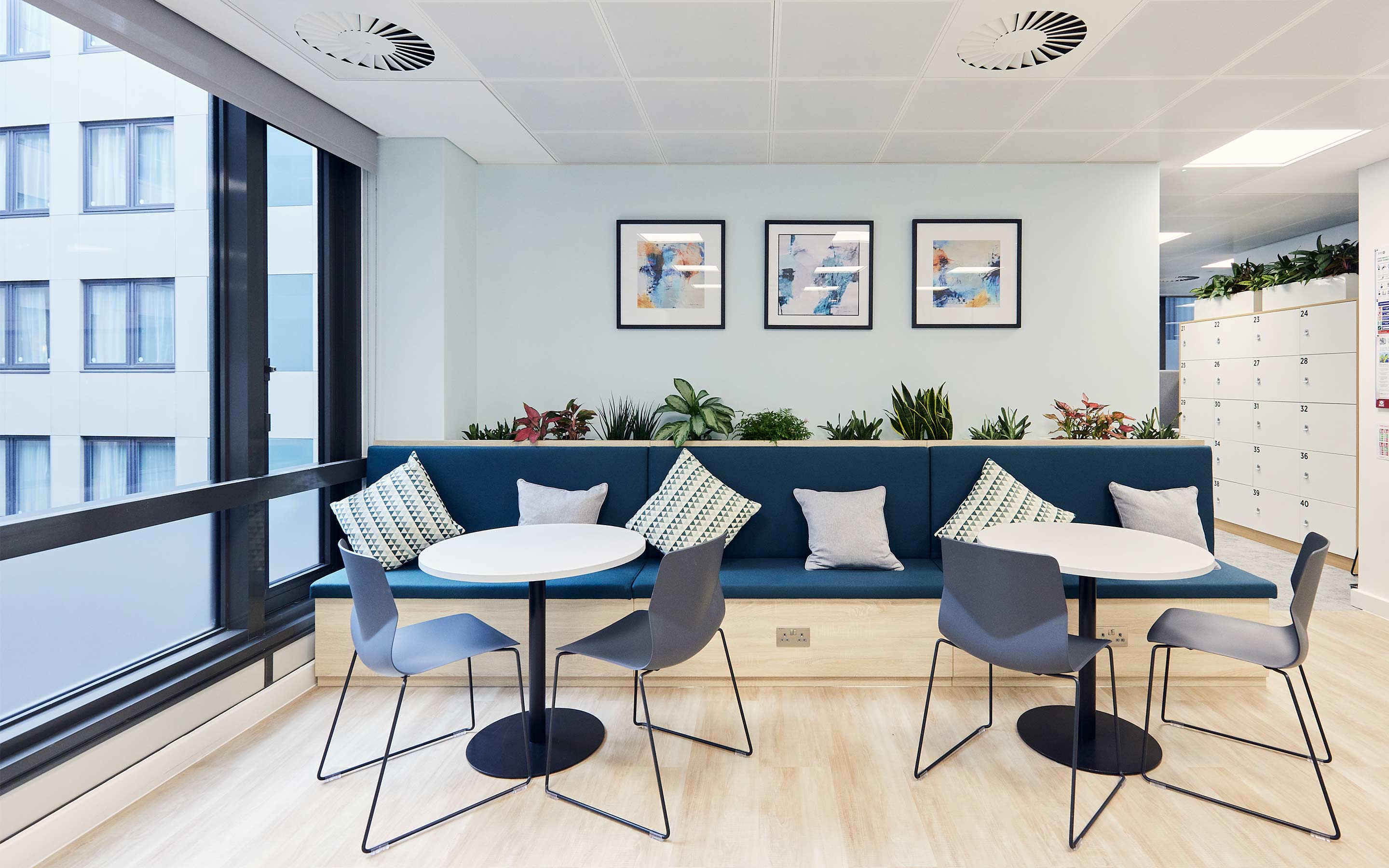 The image shows cafeteria seating in a bright office tea point. Artwork, indoor plants and light wooden flooring brightens up a banquette seating area