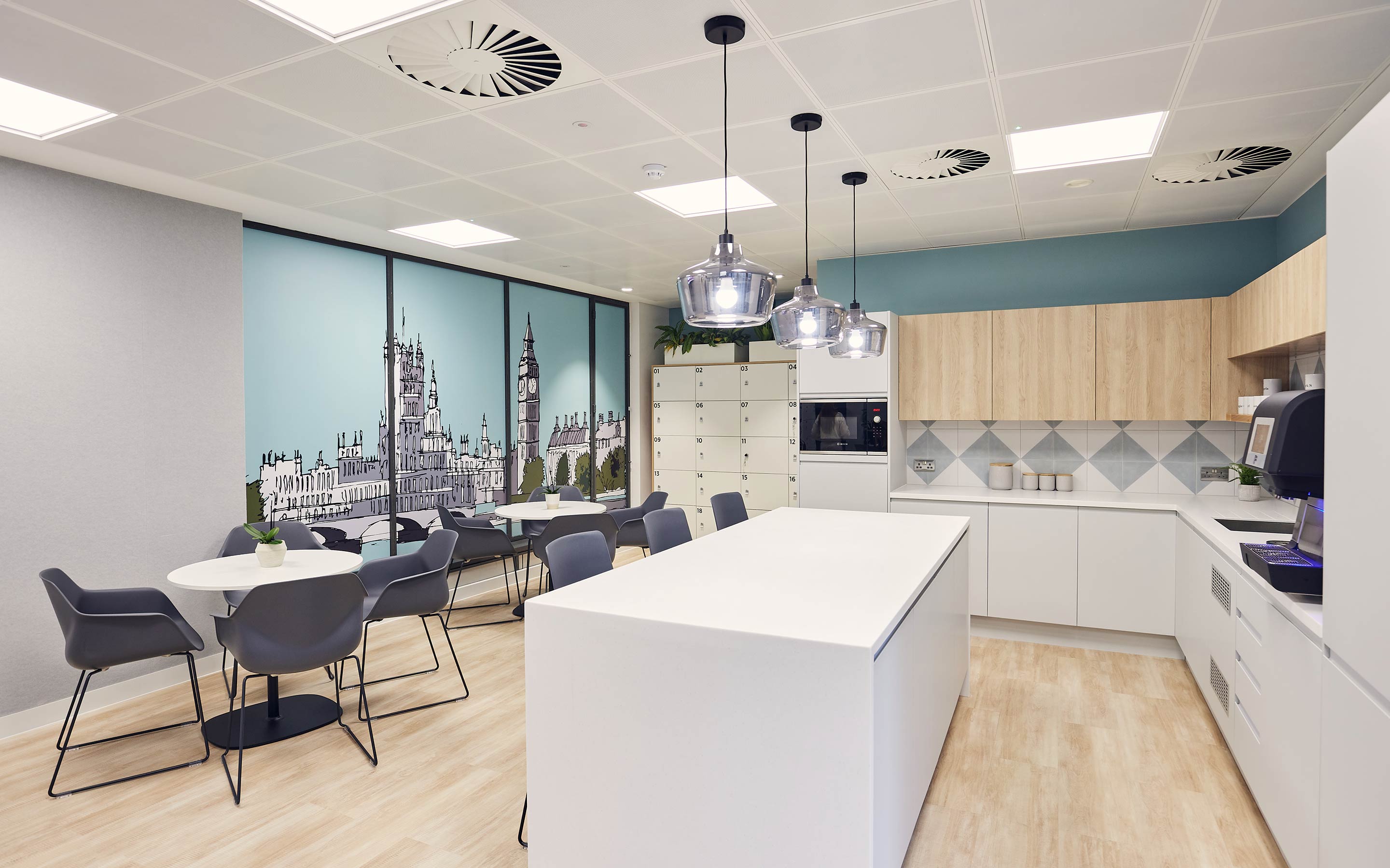 The image shows a bright, modern office tea point with kitchen facilities, and multiple seating options