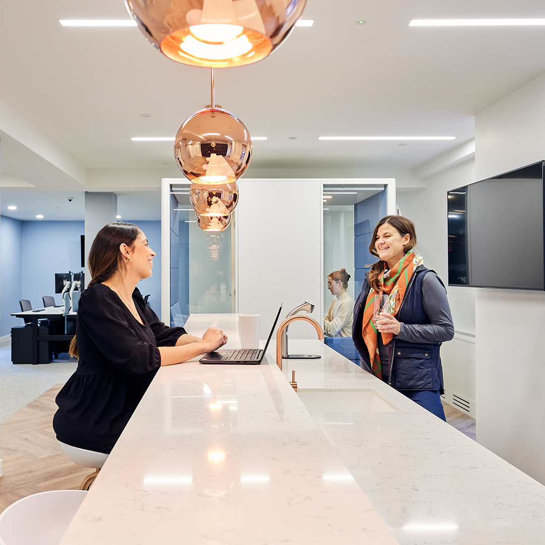 Two women laughing at a breakfast bar in an office with blue decor and large copper pendant lighting, showcasing modern office interior design