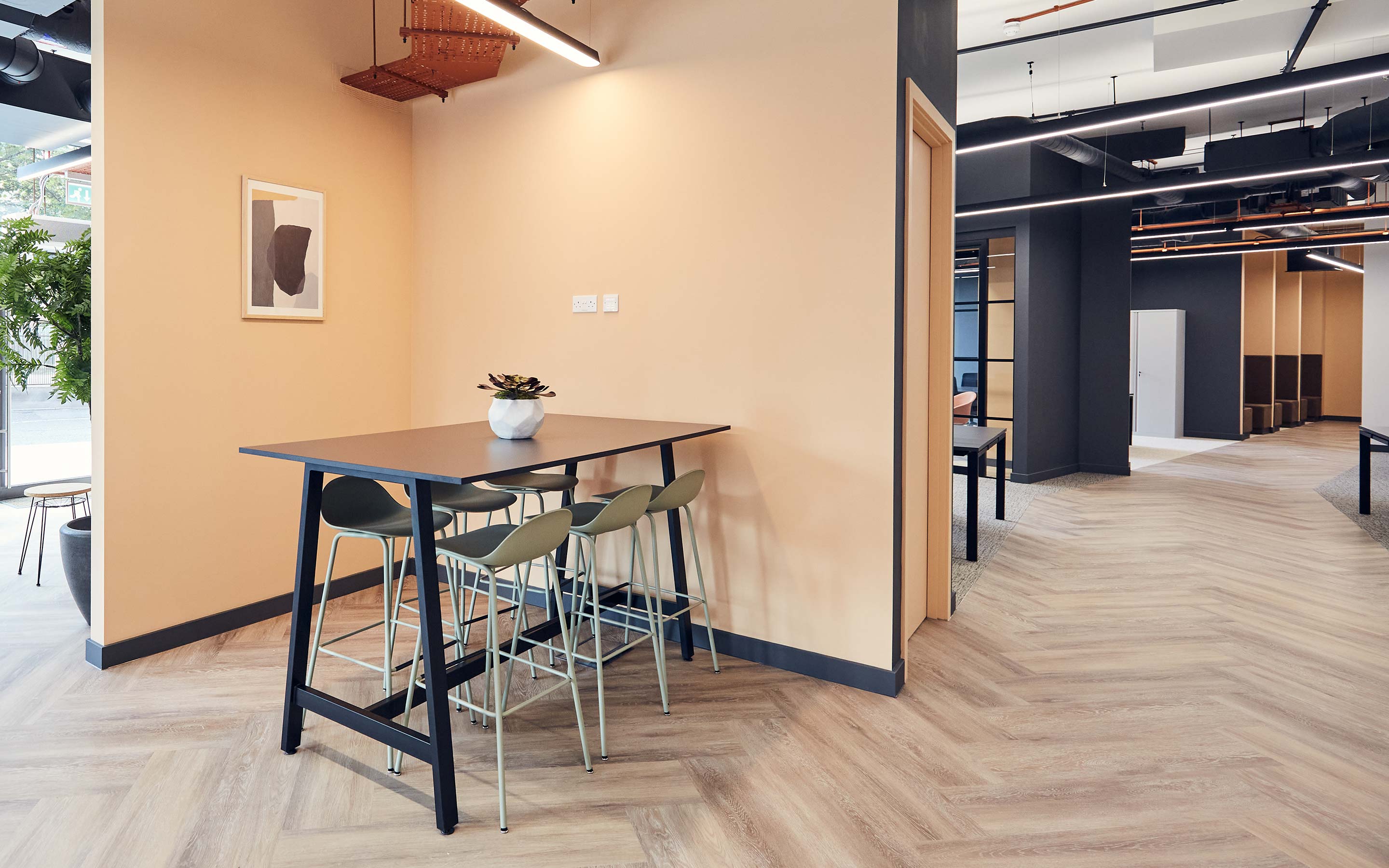 The image shows a high table and stools in an office breakout area, with peach coloured walls and wooden flooring