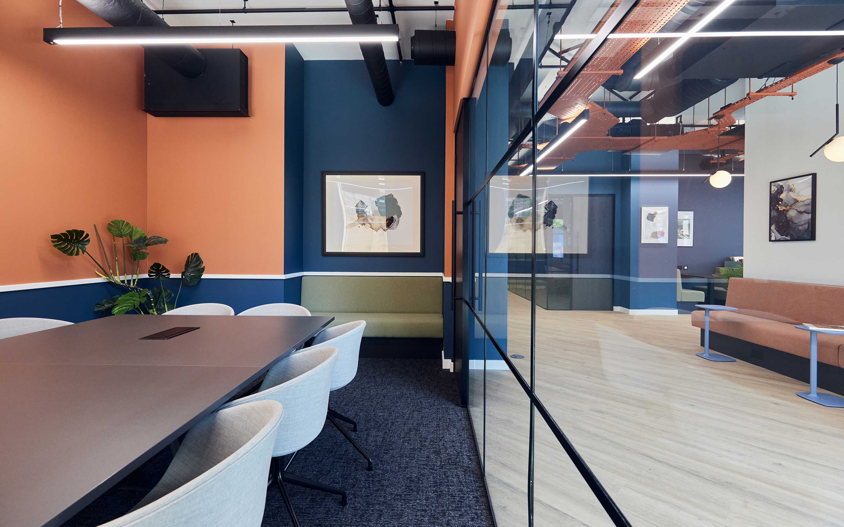 The image shows a meeting room with blue and orange walls, a large meeting table and chairs, and a large glass door with views of the rest of the office