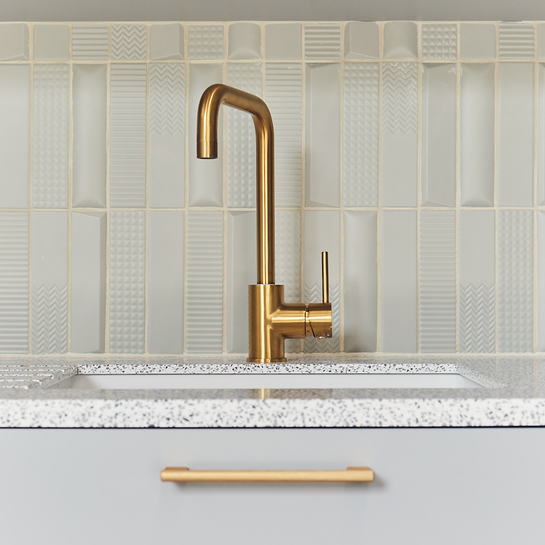 A gold tap set against pale grey backsplash kitchen tiles in an office teapoint