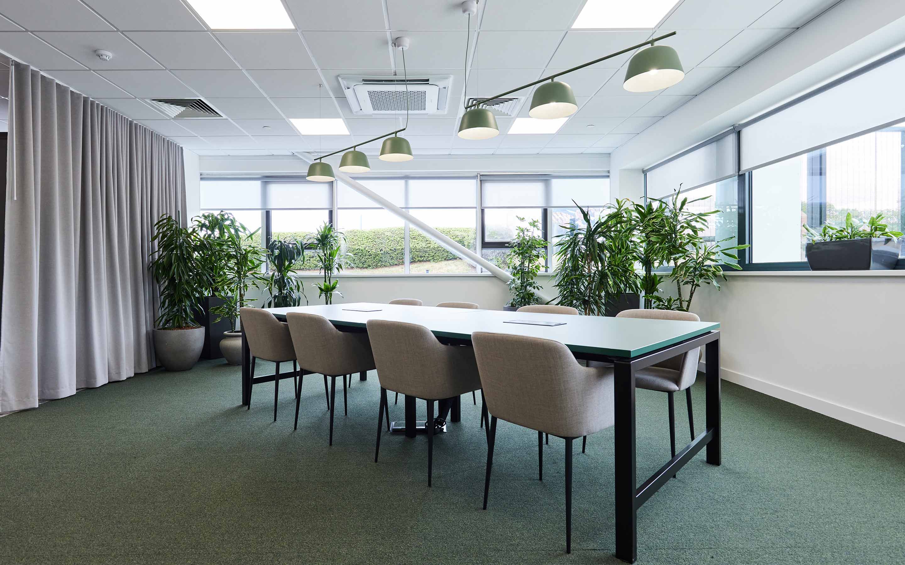 An office desking area features pendant lighting, soft grey office chairs, and indoor planting