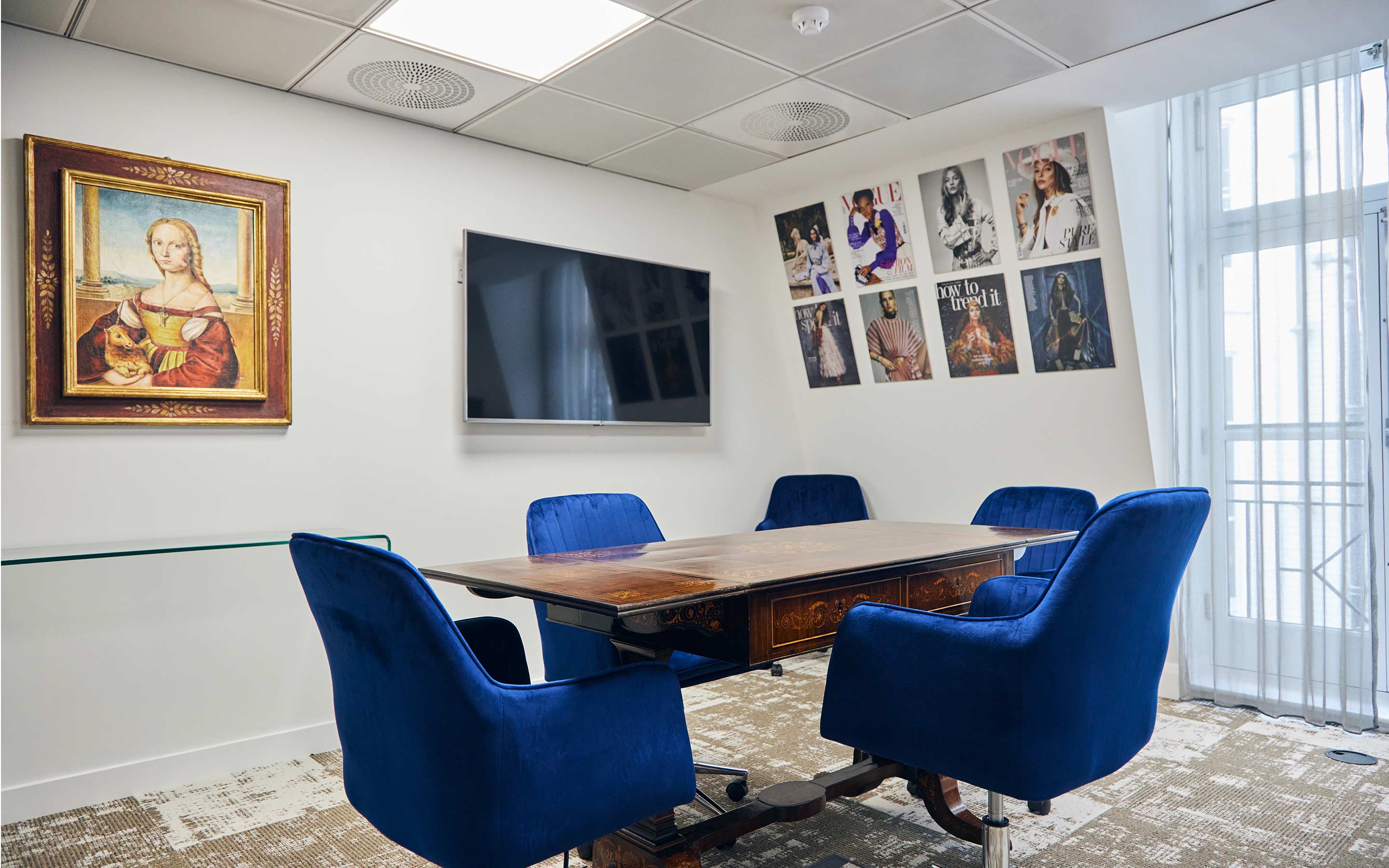 The image shows a bespoke office meeting room with artwork, and magazine issues hung on the walls, and plush blue meeting chairs
