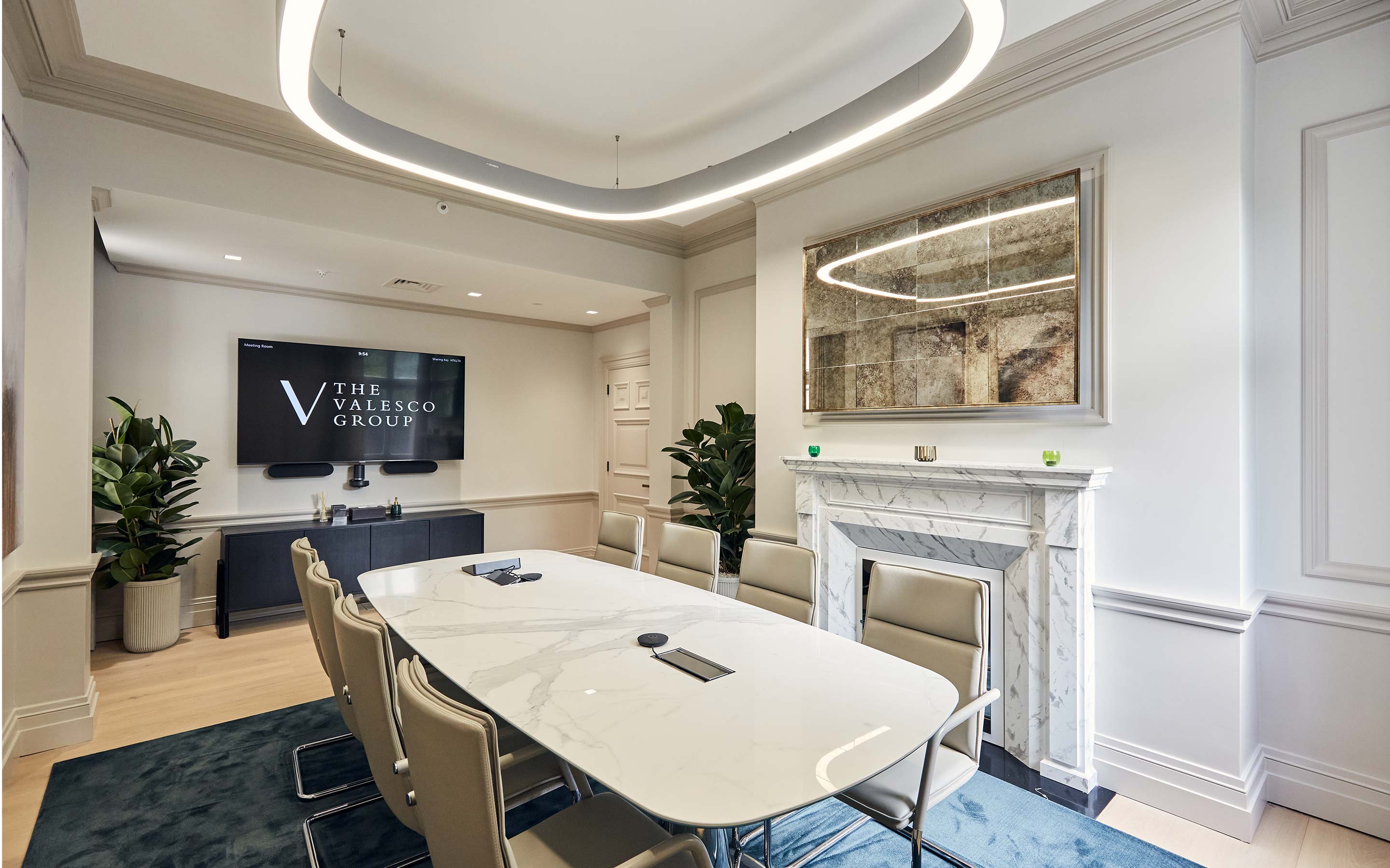 White marble table and white leather chairs in a meeting room, with a marble fireplace, tall plants in pots in the corners of the room, and a large television screen