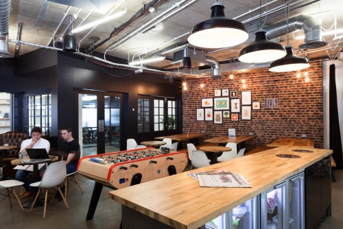 A dark social and kitchen space with soft amber lighting and an exposed brick wall at one end a foosball table in the centre and wooden tables with benches or white seats surrounding