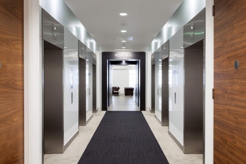 A corridor with three metal lifts on either side of the wall  a grey striped carpet running down the centre of the floor and white carpet on either side