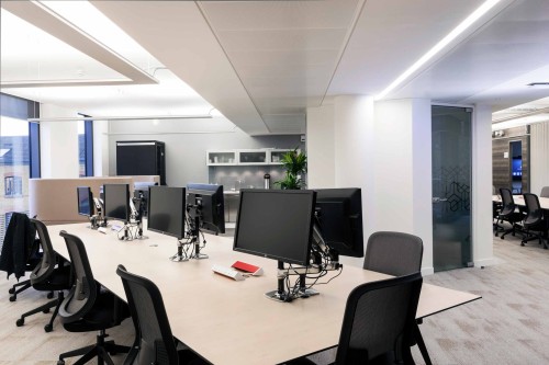 A large rectangular light wood finished desk in the centre of a white workspace with black chairs and monitors